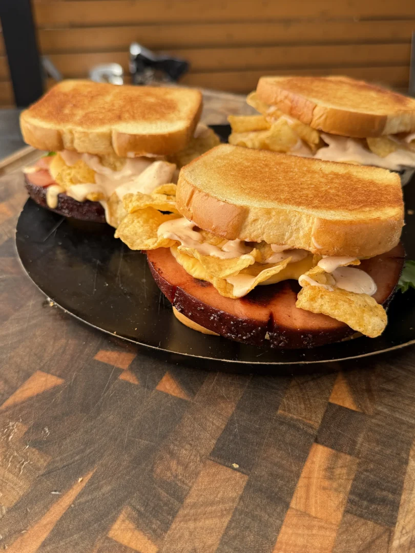 Smoked and fried bologna sandwiches on toasted bread with potato chips and sauce on a dark round platter