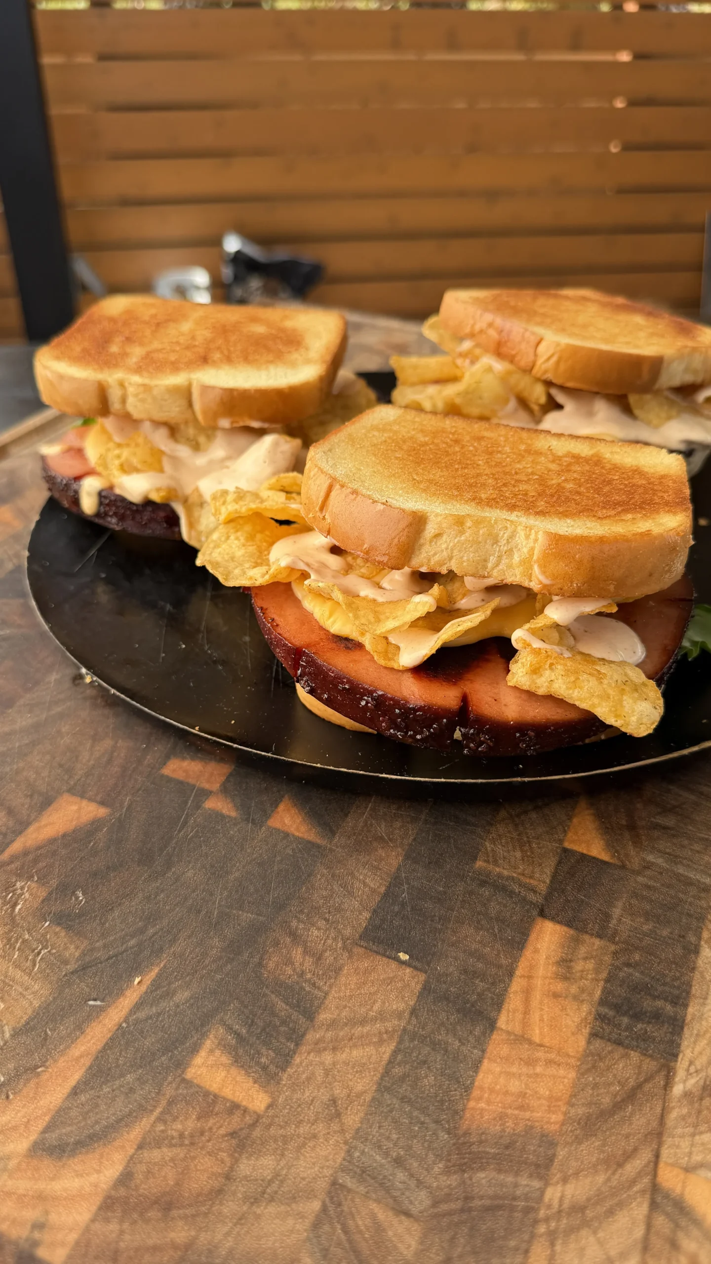 Smoked and fried bologna sandwiches on toasted bread with potato chips and sauce on a dark round platter