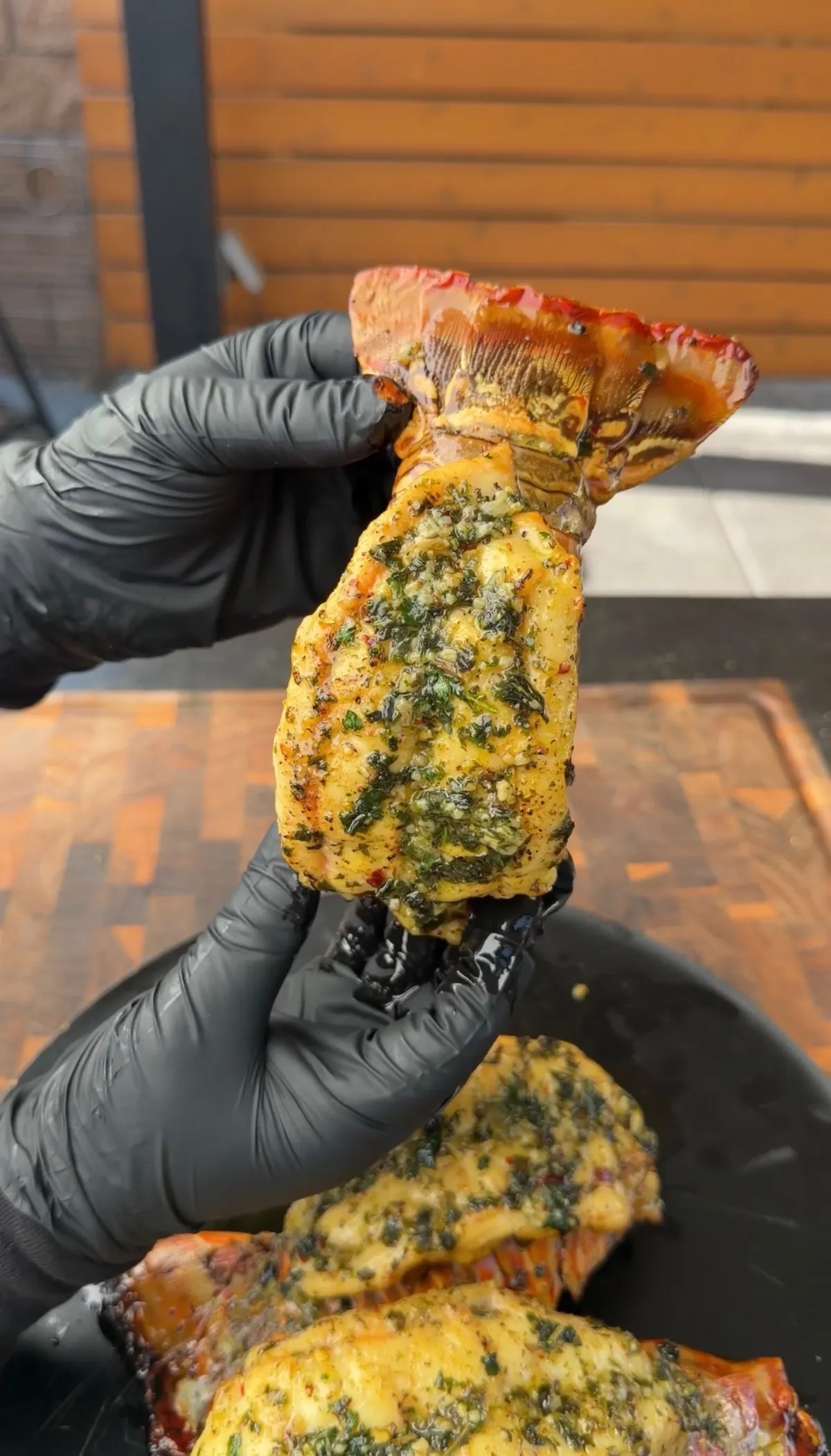 Hands in black gloves holding a smoked lobster tail coated in garlic herb butter over a cutting board