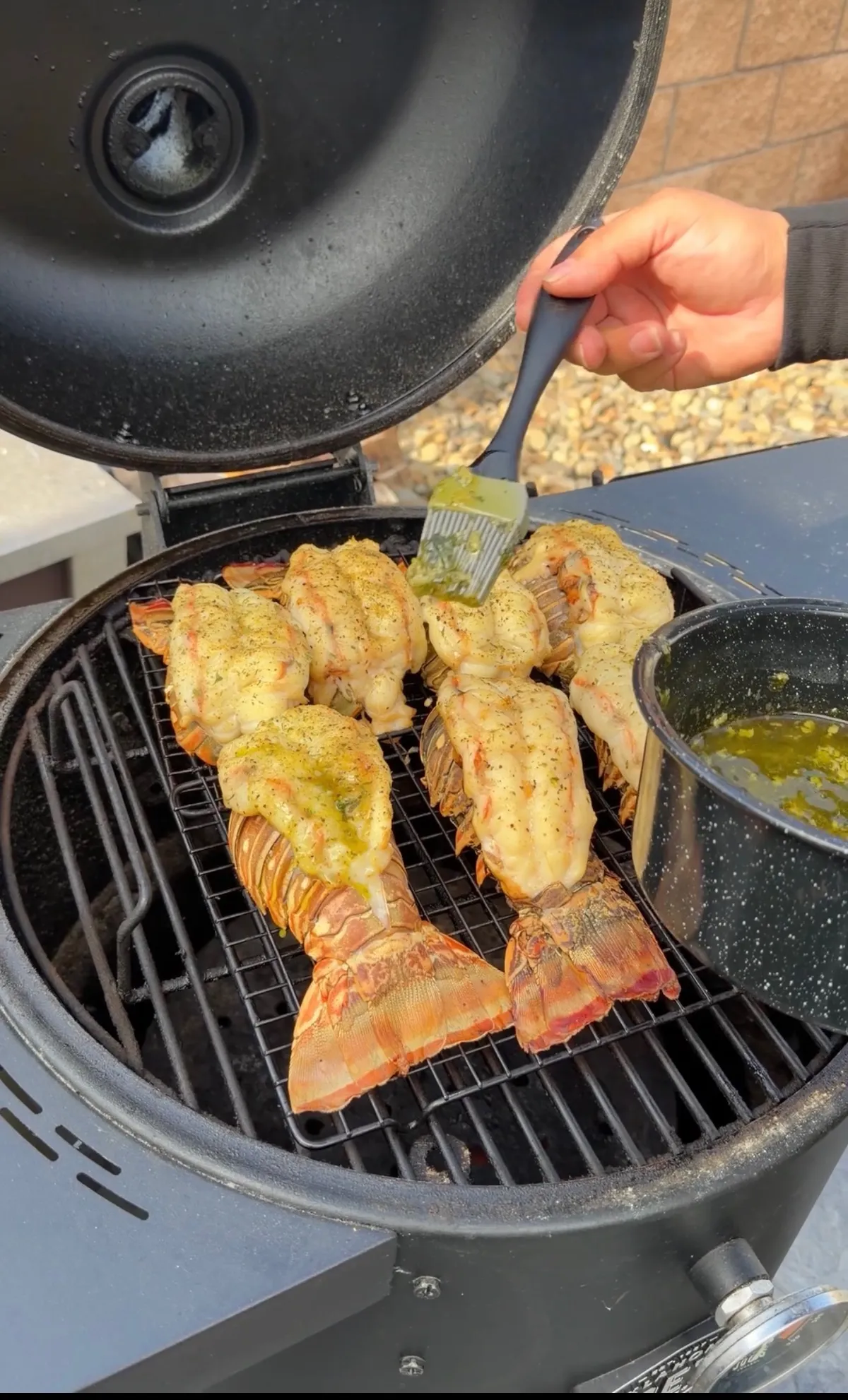 Lobster tails grilling on a kamado smoker being basted with herb garlic butter using a silicone brush