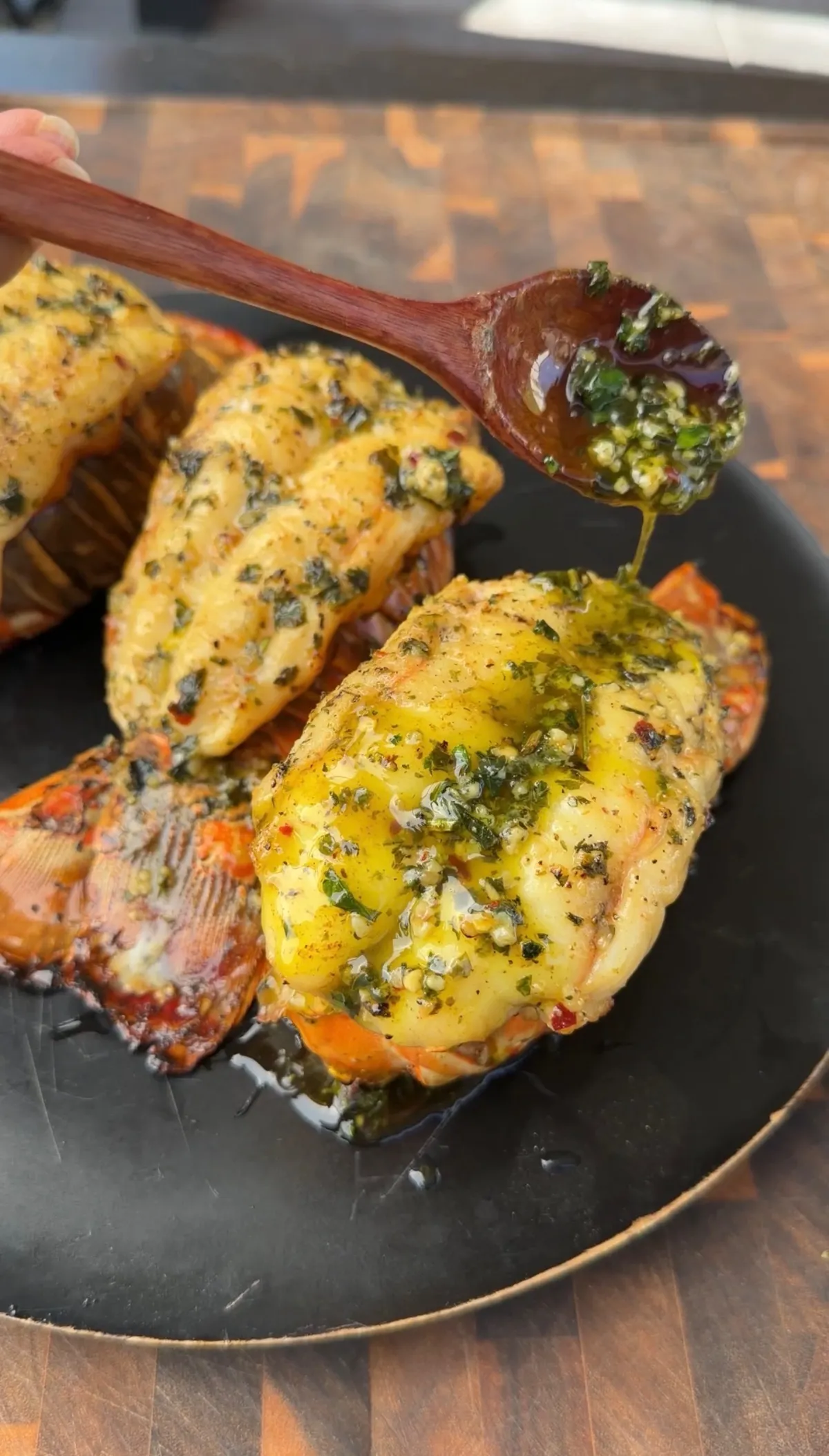 Smoked lobster tails on black plate being basted with garlic herb butter sauce using a wooden spoon