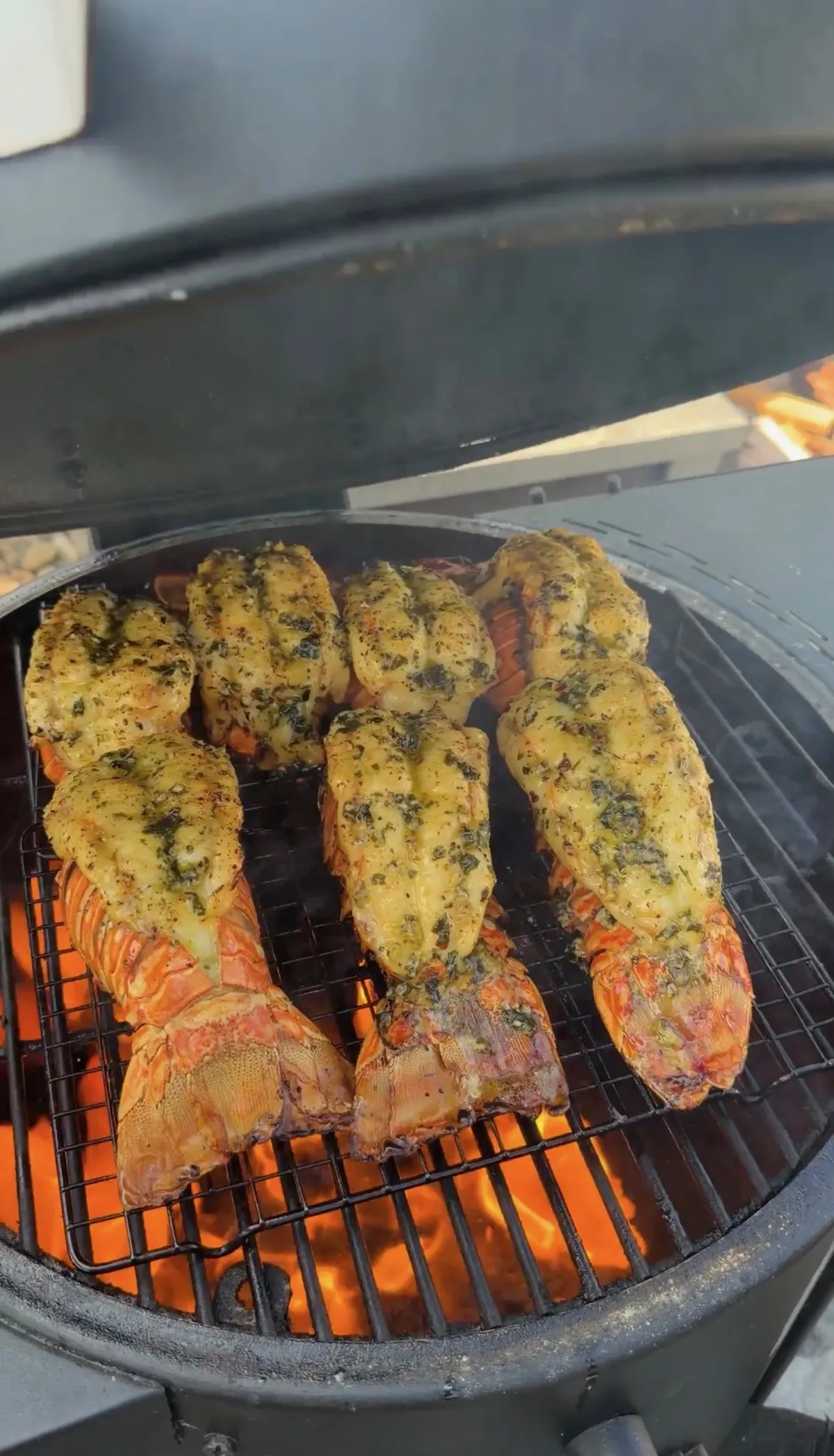 Six lobster tails topped with herb garlic butter grilling over hot charcoal on a round kamado-style smoker