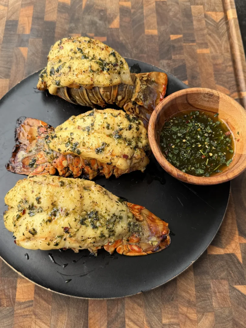 Three smoked lobster tails coated in herb butter on a black plate with a wooden bowl of chimichurri sauce