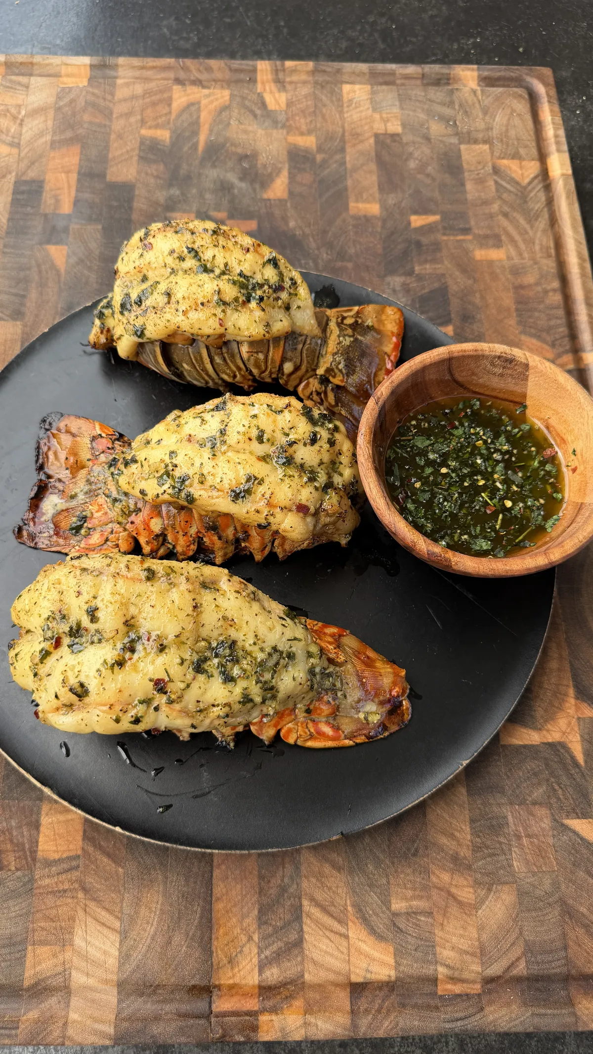 Three smoked lobster tails coated in herb butter on a black plate with a wooden bowl of chimichurri sauce