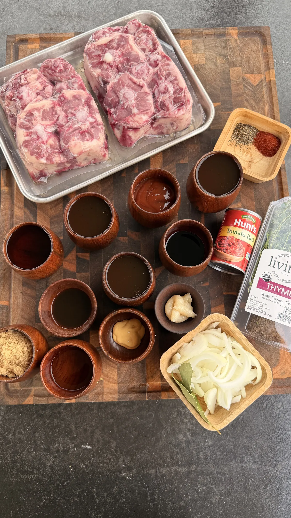 Raw oxtails with bourbon, beef broth, BBQ sauce, tomato paste, Dijon mustard, onion, thyme, and spices on a cutting board