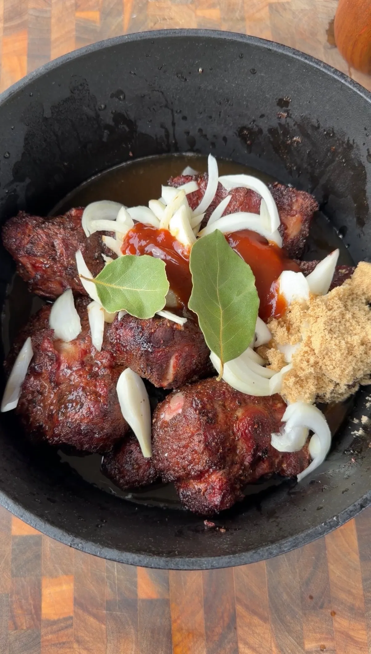 Smoked oxtails in a Dutch oven with sliced onion, garlic, BBQ sauce, brown sugar, and bay leaves before braising
