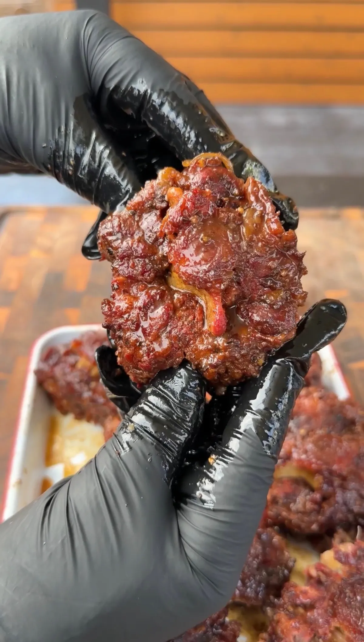 Gloved hands holding a saucy, fork-tender smoked oxtail piece over a pan of brown sugar bourbon braised oxtails