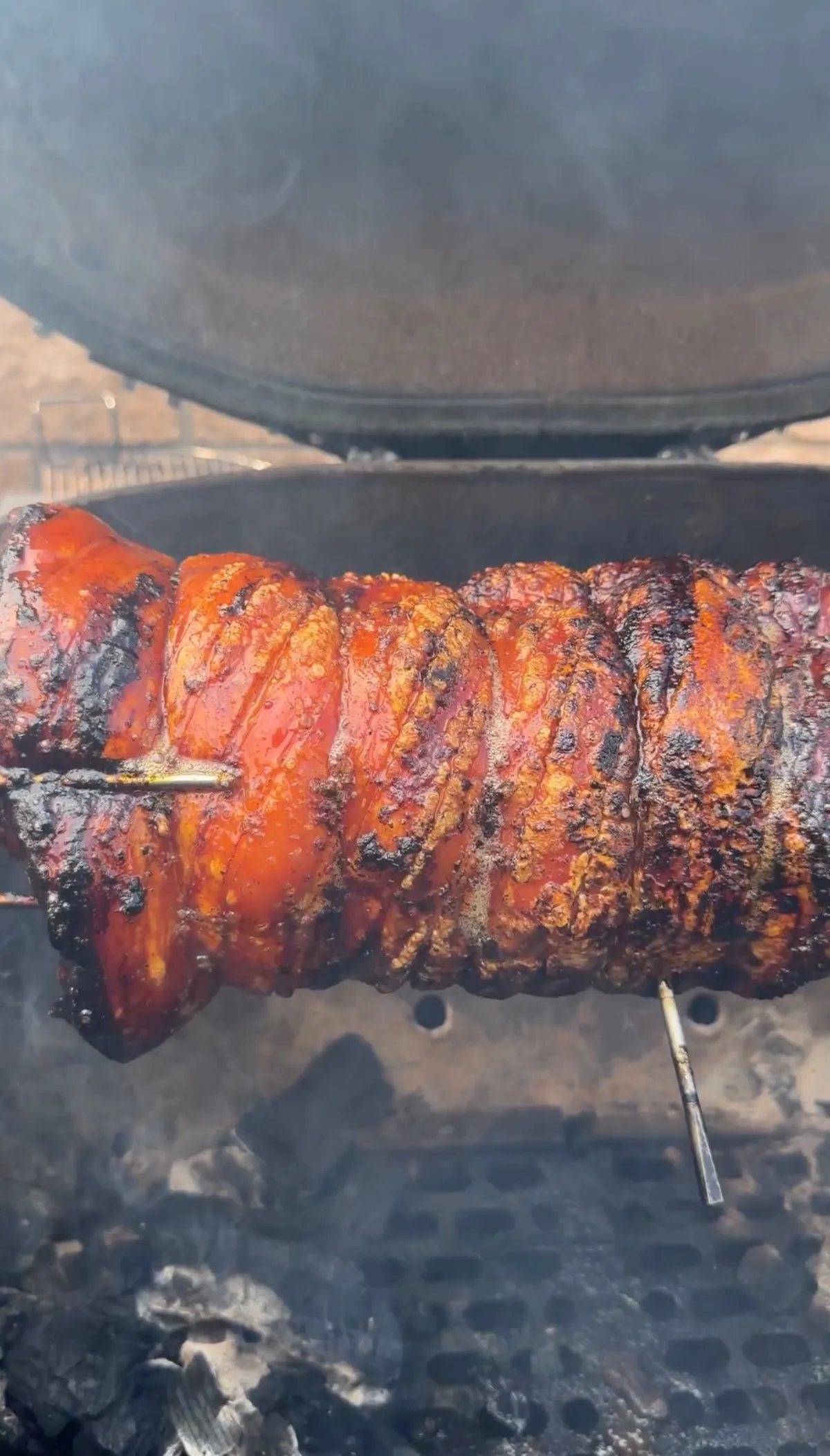 Smoky pork belly porchetta roll with caramelized crust cooking on a rotisserie spit over charcoal