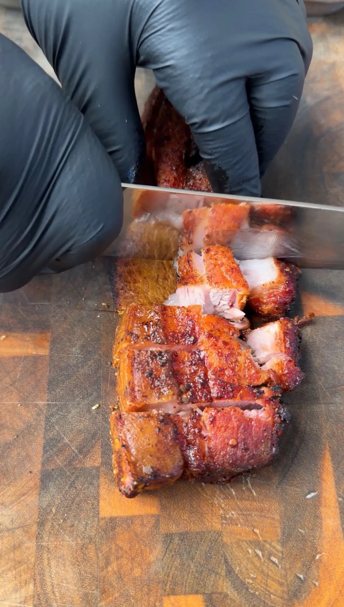 Hands in black gloves slicing crispy smoked pork belly into cubes on a wooden cutting board for chicharron tacos