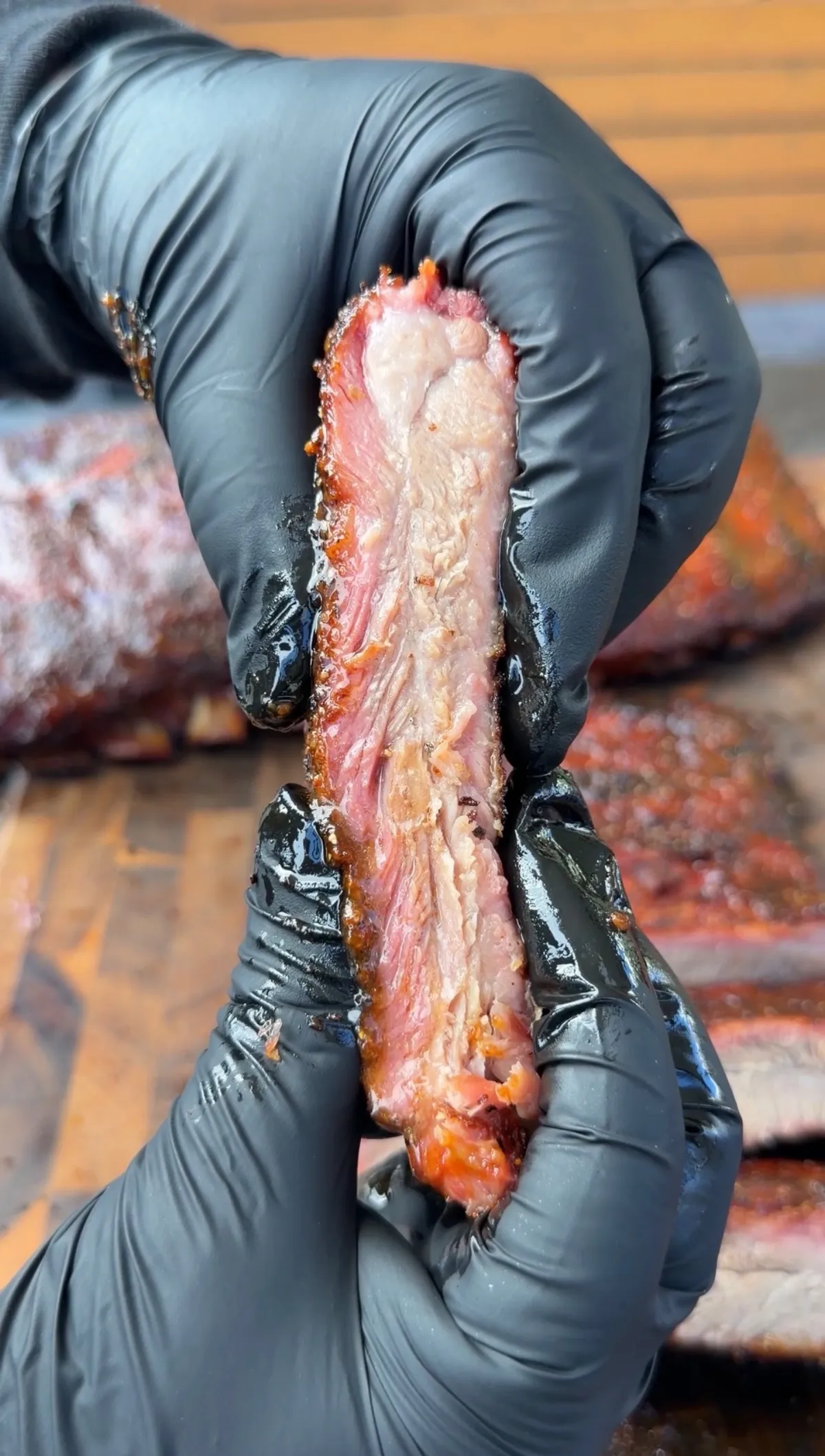 Hands in black gloves holding a single smoked rib showing juicy pink smoke ring and tender meat interior