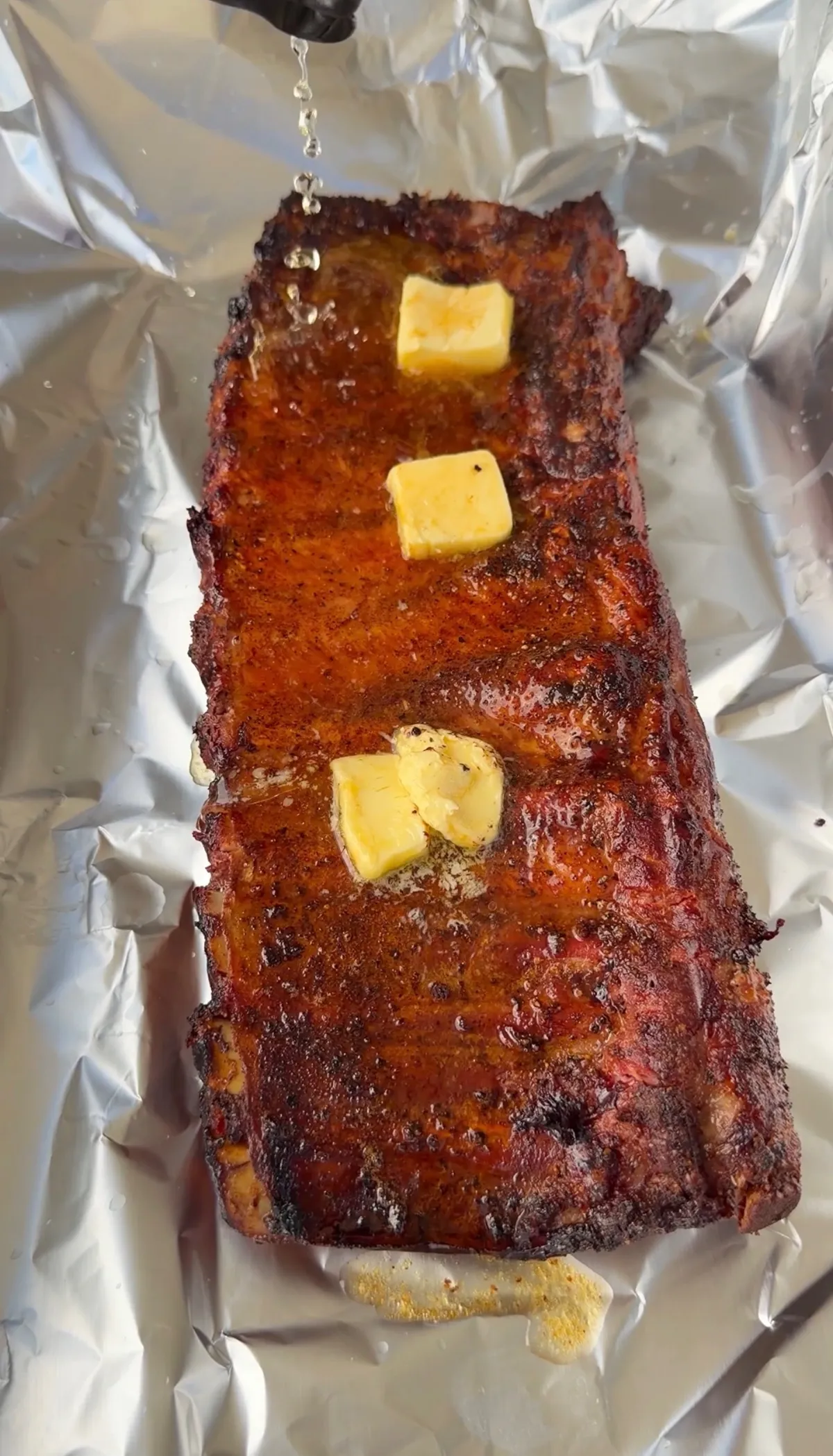 Smoked ribs rack with melting butter pats on aluminum foil, glazed with a rich spice rub crust