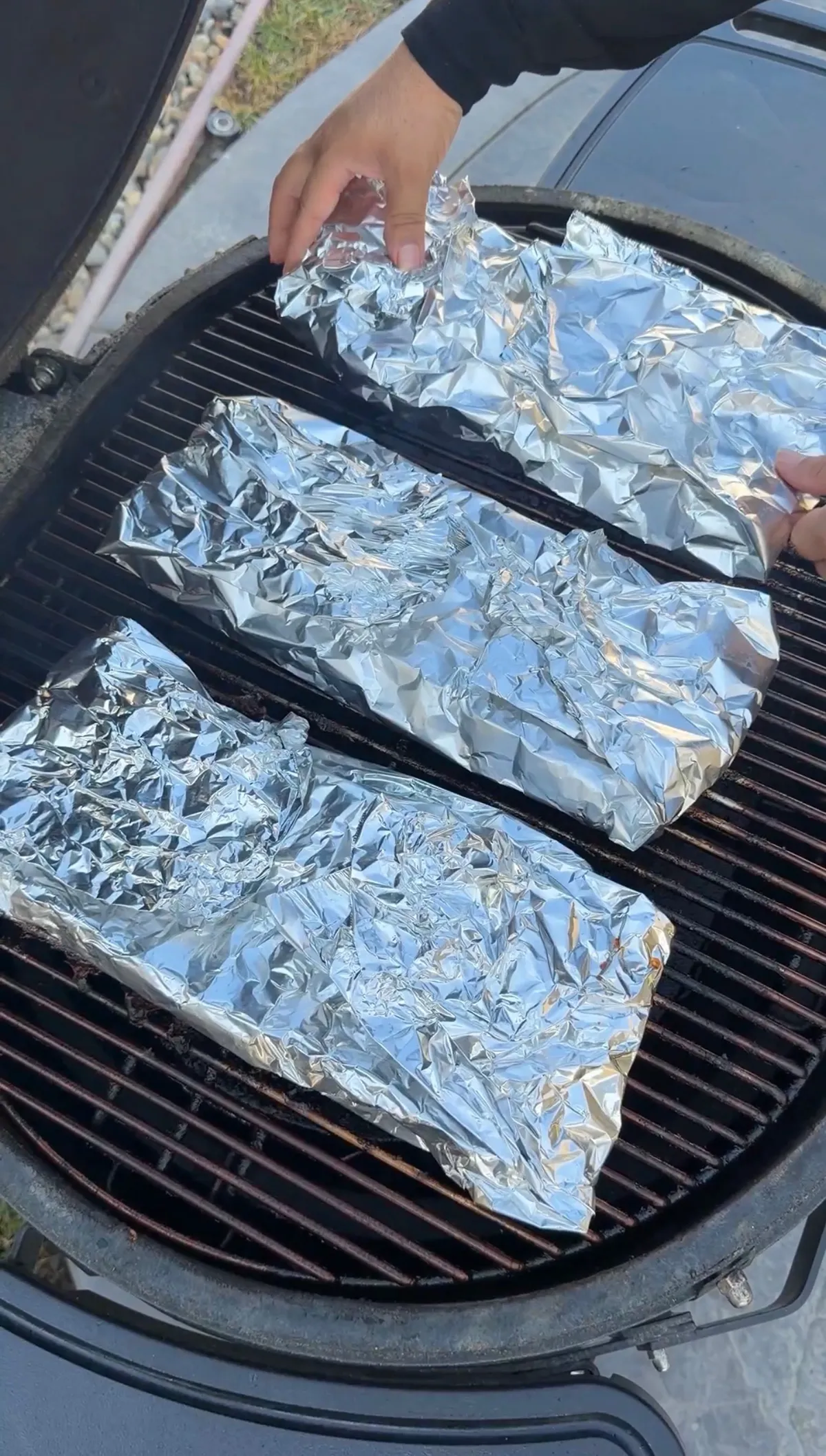 Three racks of ribs wrapped in aluminum foil packets placed on a charcoal grill grate