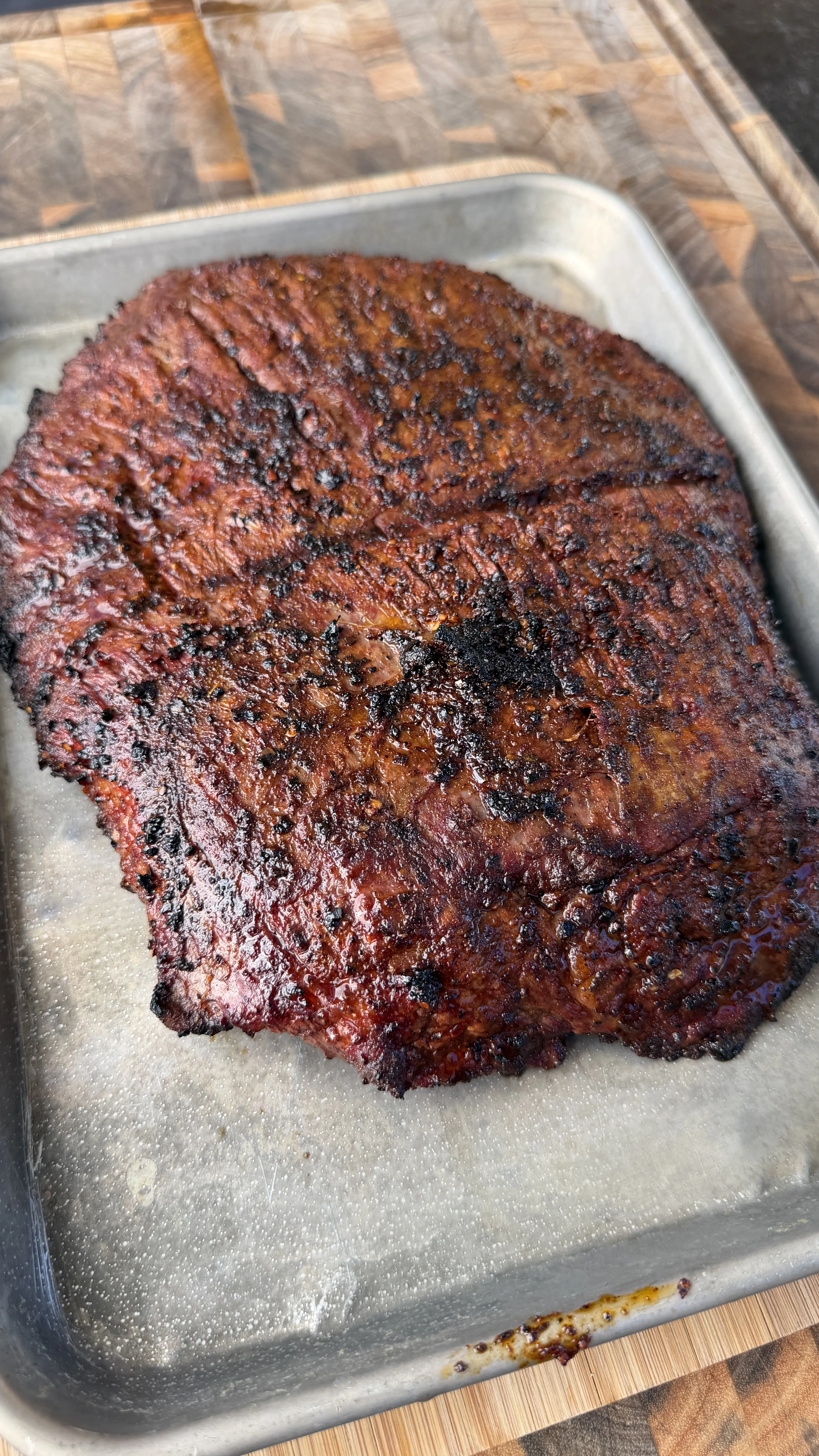 Charred carne asada with dark bark resting on a silver sheet pan over a wooden cutting board