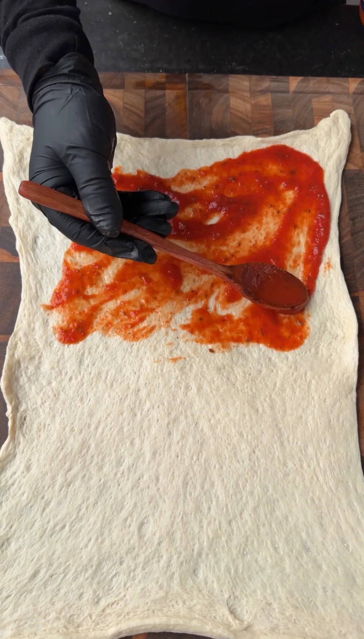 Gloved hand spreading marinara sauce over half of a rolled-out Pillsbury pizza dough rectangle on a wooden board