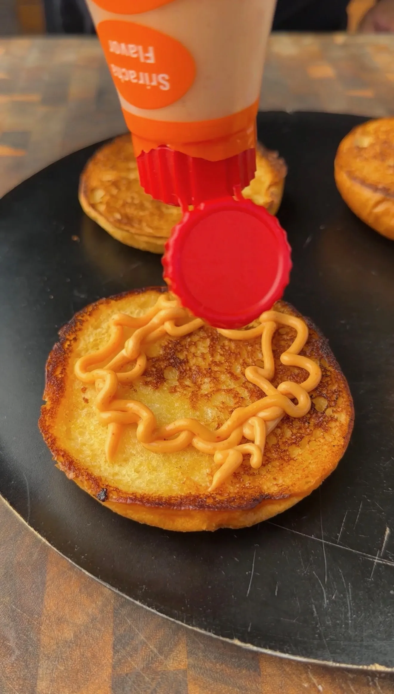 Toasted burger bun on a black plate being drizzled with Sriracha mayo from a squeeze bottle