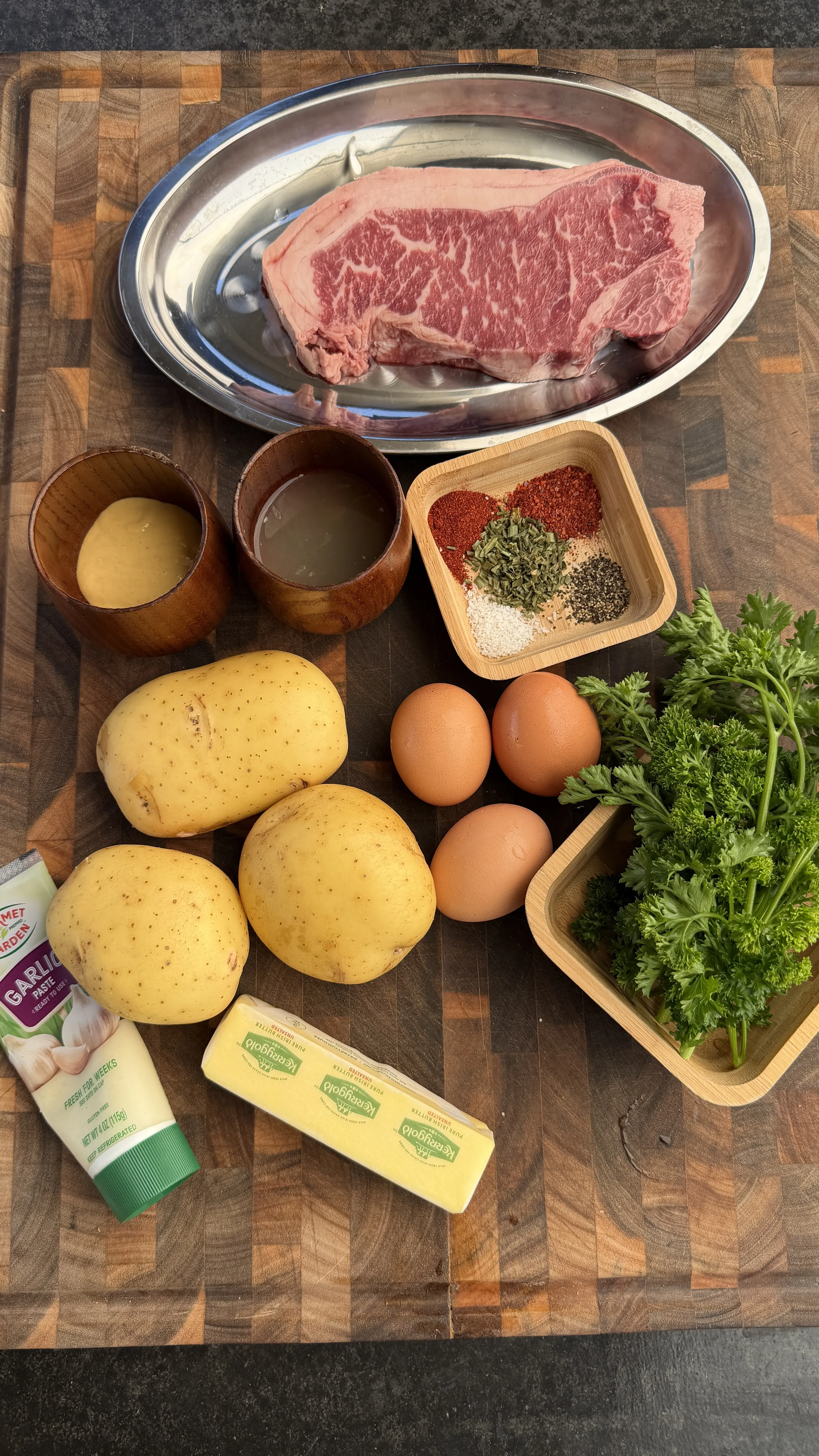 Raw ingredients for steak, eggs, and crispy potatoes with cowboy butter, including steak, Yukon Gold potatoes, eggs, butter, garlic, spices, and fresh parsley on a wooden cutting board