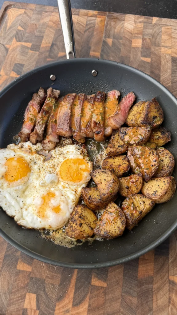 Sliced medium-rare steak, three fried eggs, and crispy roasted potatoes in a black skillet with cowboy butter sauce