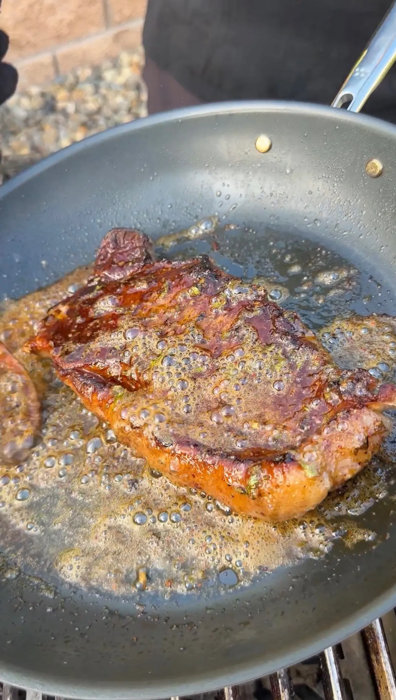 Seasoned steak searing in a bubbling cowboy butter sauce in a dark skillet outdoors