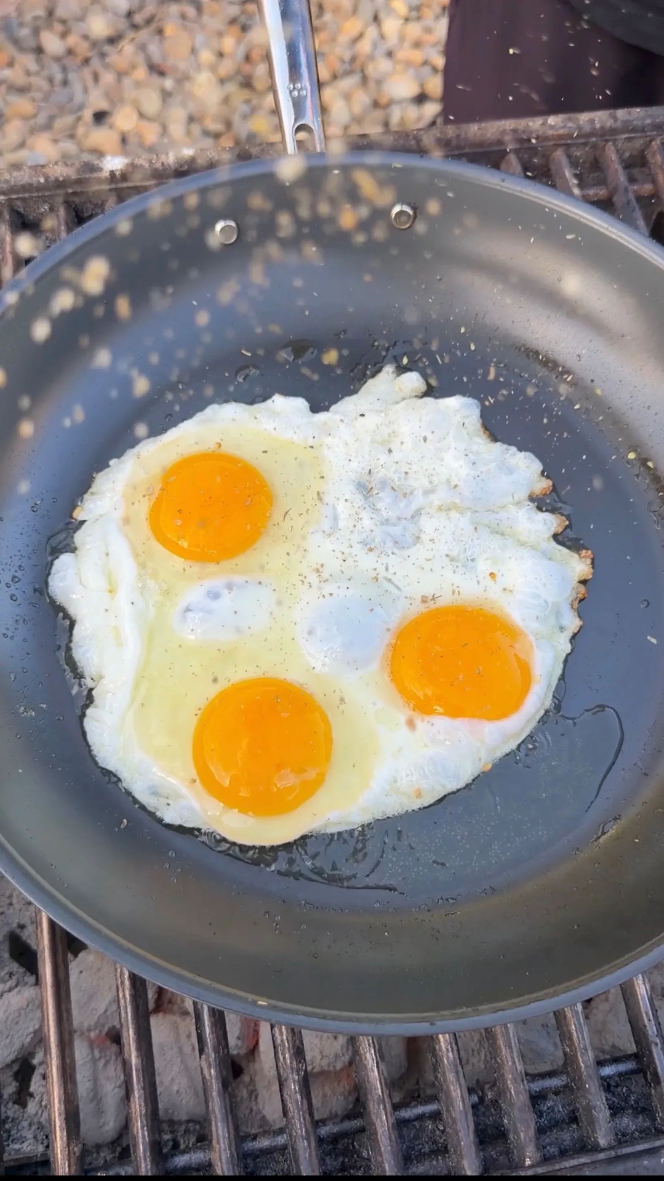 Three sunny-side-up eggs with bright orange yolks frying in a skillet on an outdoor grill grate