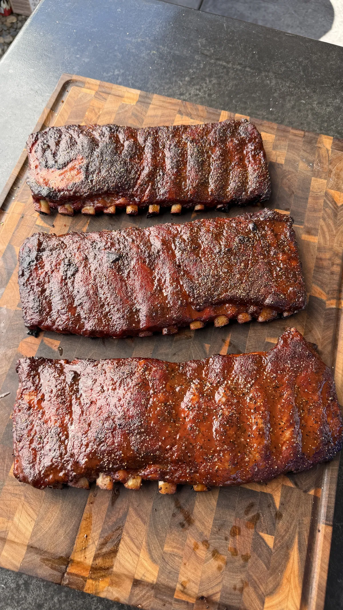 Three racks of smoked BBQ ribs with dark bark and glaze resting on a wooden cutting board