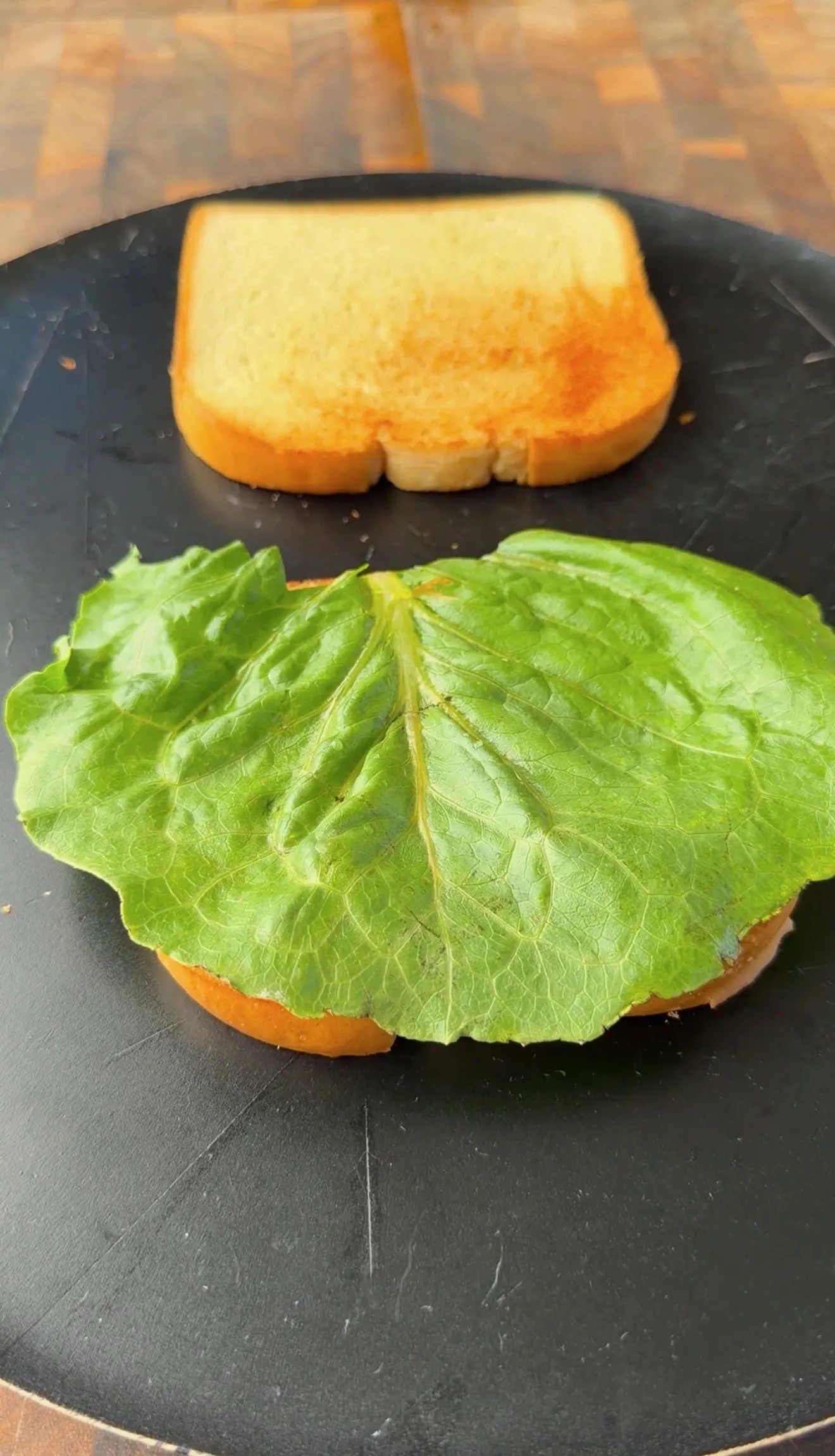 Toasted bread slices with a large fresh lettuce leaf on a black plate, prepping a smoked fried bologna sandwich