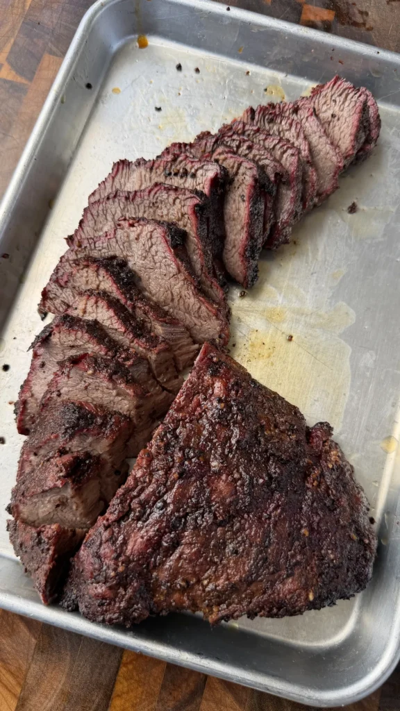 Sliced trisket (tri-tip cooked like brisket) with dark bark and pink smoke ring on a metal tray