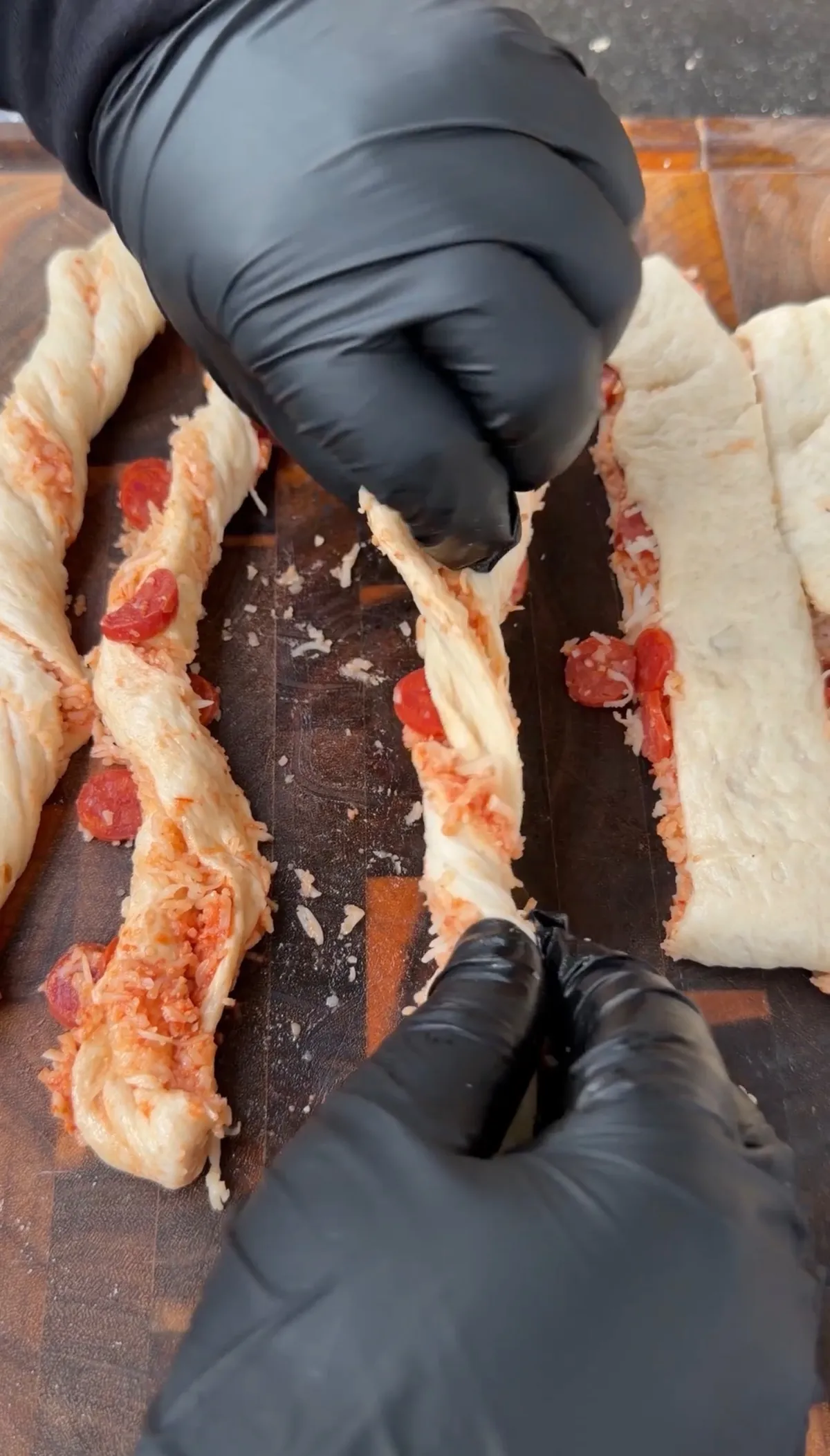Gloved hands twisting strips of Pillsbury pizza dough filled with marinara, mozzarella, Parmesan, and mini pepperonis on a wooden cutting board