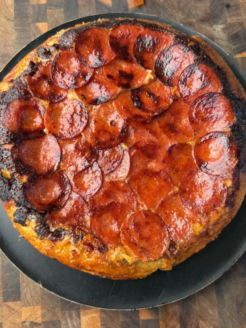 Upside down pepperoni pizza with caramelized crispy pepperoni on top, served on a black round pan over a wood board