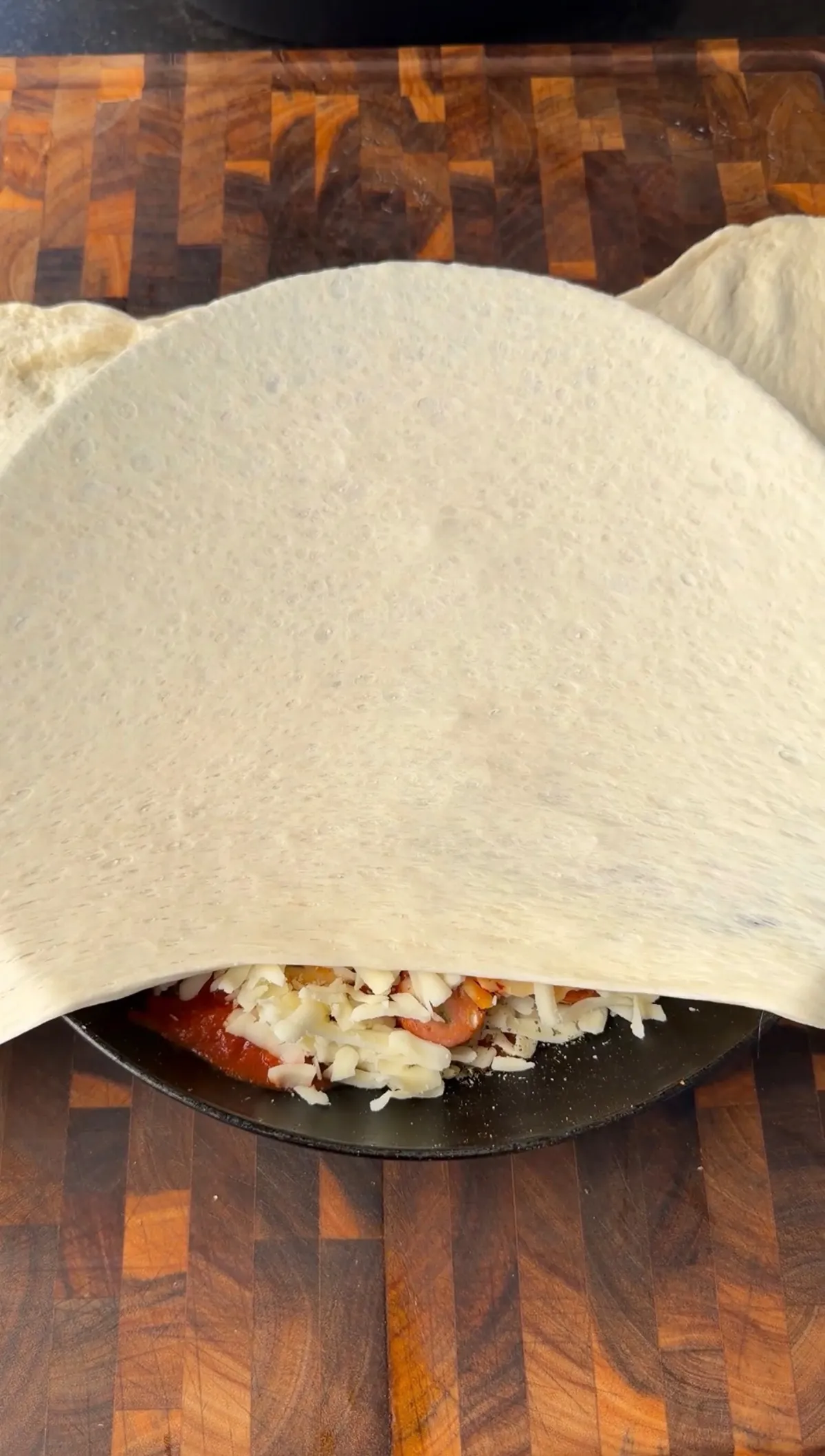 Pizza dough being folded over tomato sauce, shredded cheese, and toppings in a dark skillet for upside down pizza