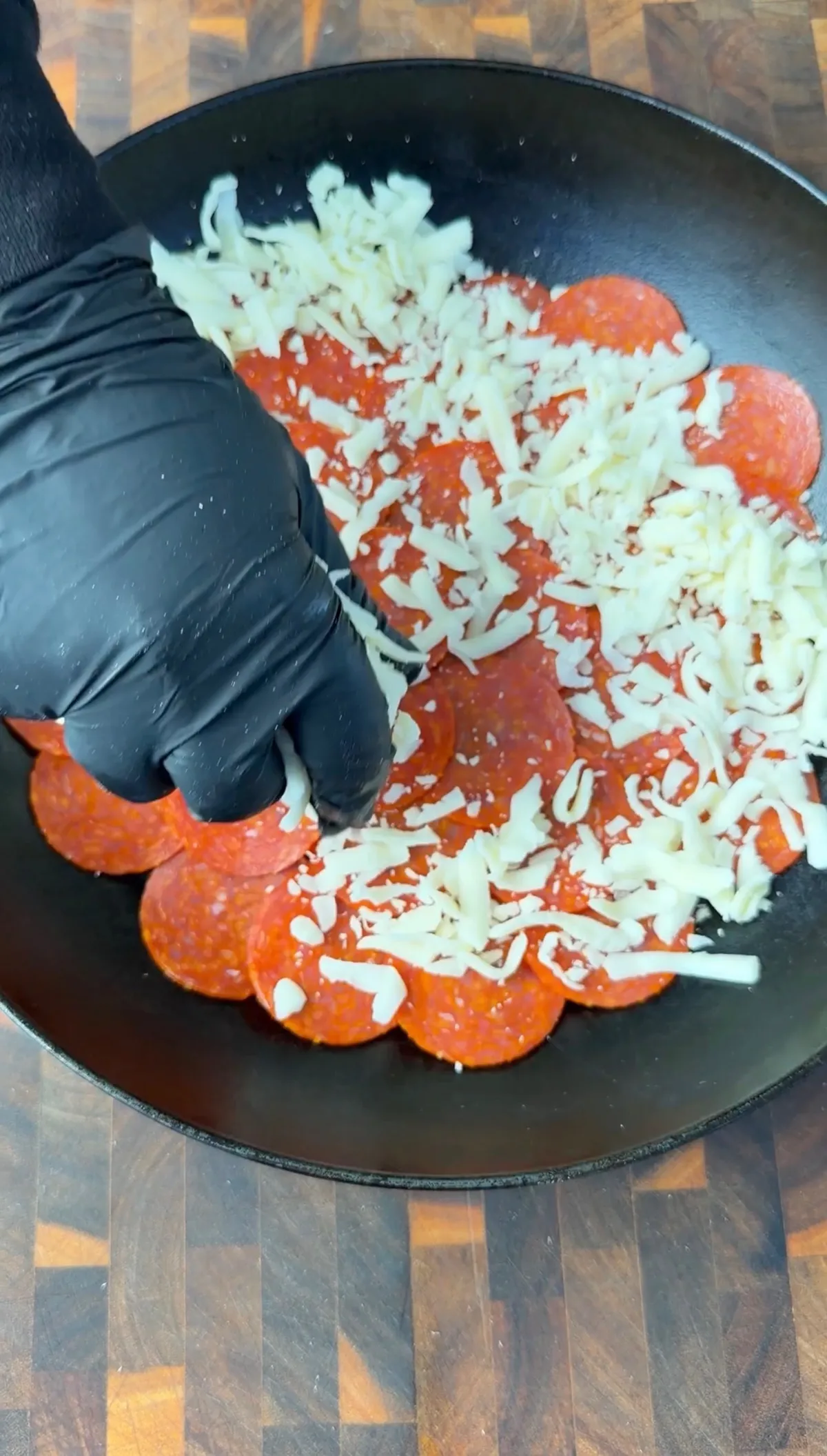 Black-gloved hand sprinkling shredded mozzarella over pepperoni slices in a cast iron skillet