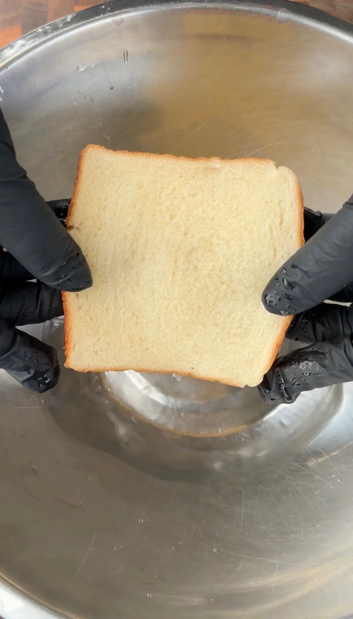 Gloved hands holding a slice of white sandwich bread over a stainless steel bowl for potato cheese balls recipe