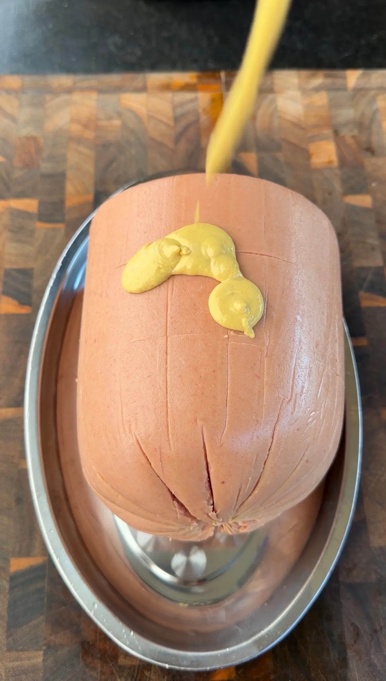 Large whole bologna scored with crosshatch cuts and yellow mustard drizzled on top in a silver roasting pan