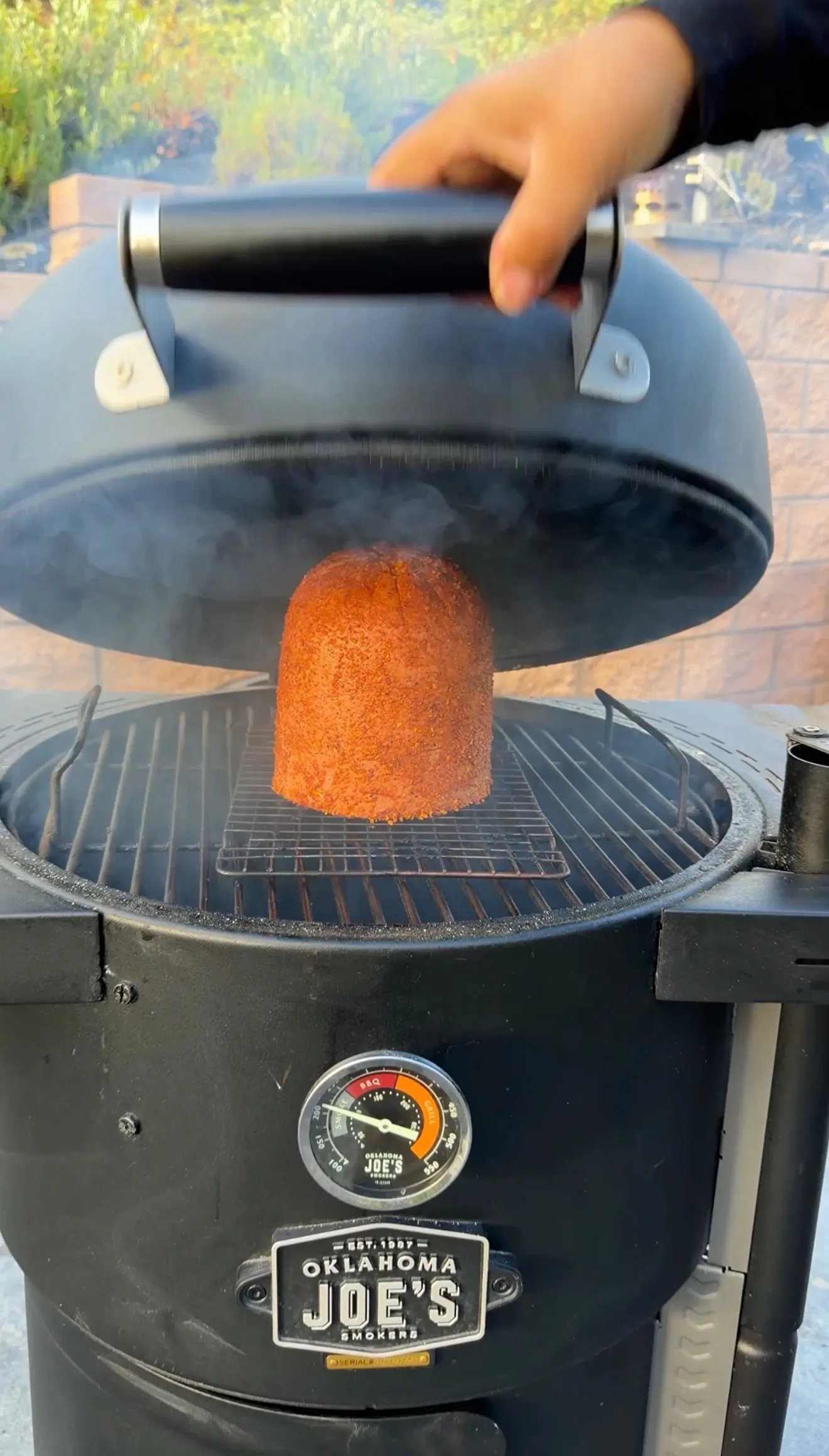 A large whole bologna covered in red seasoning rub smoking on an Oklahoma Joe's charcoal smoker with lid lifted