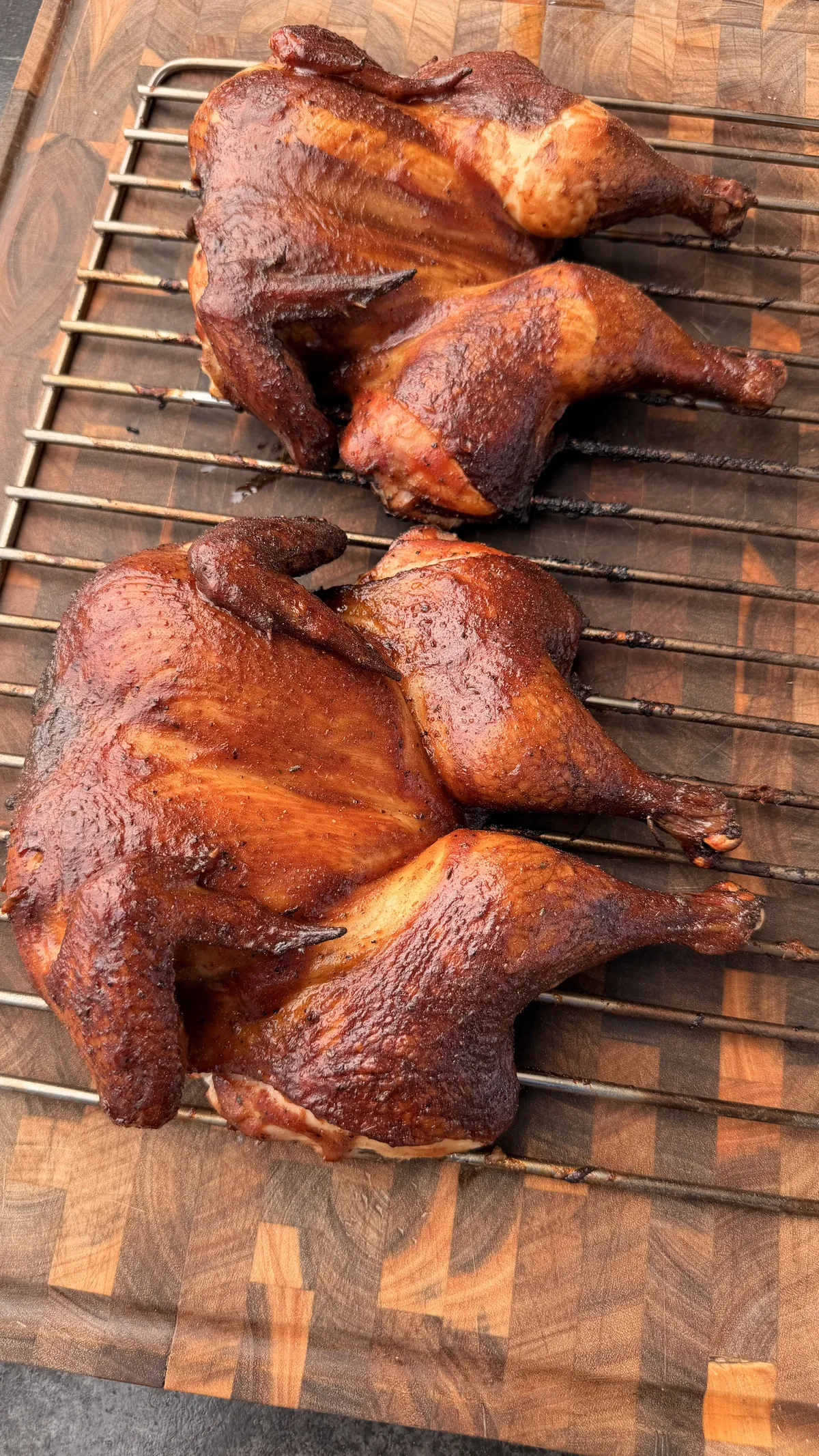 Two BBQ-glazed applewood smoked spatchcock chickens resting on a wire rack over a wooden cutting board