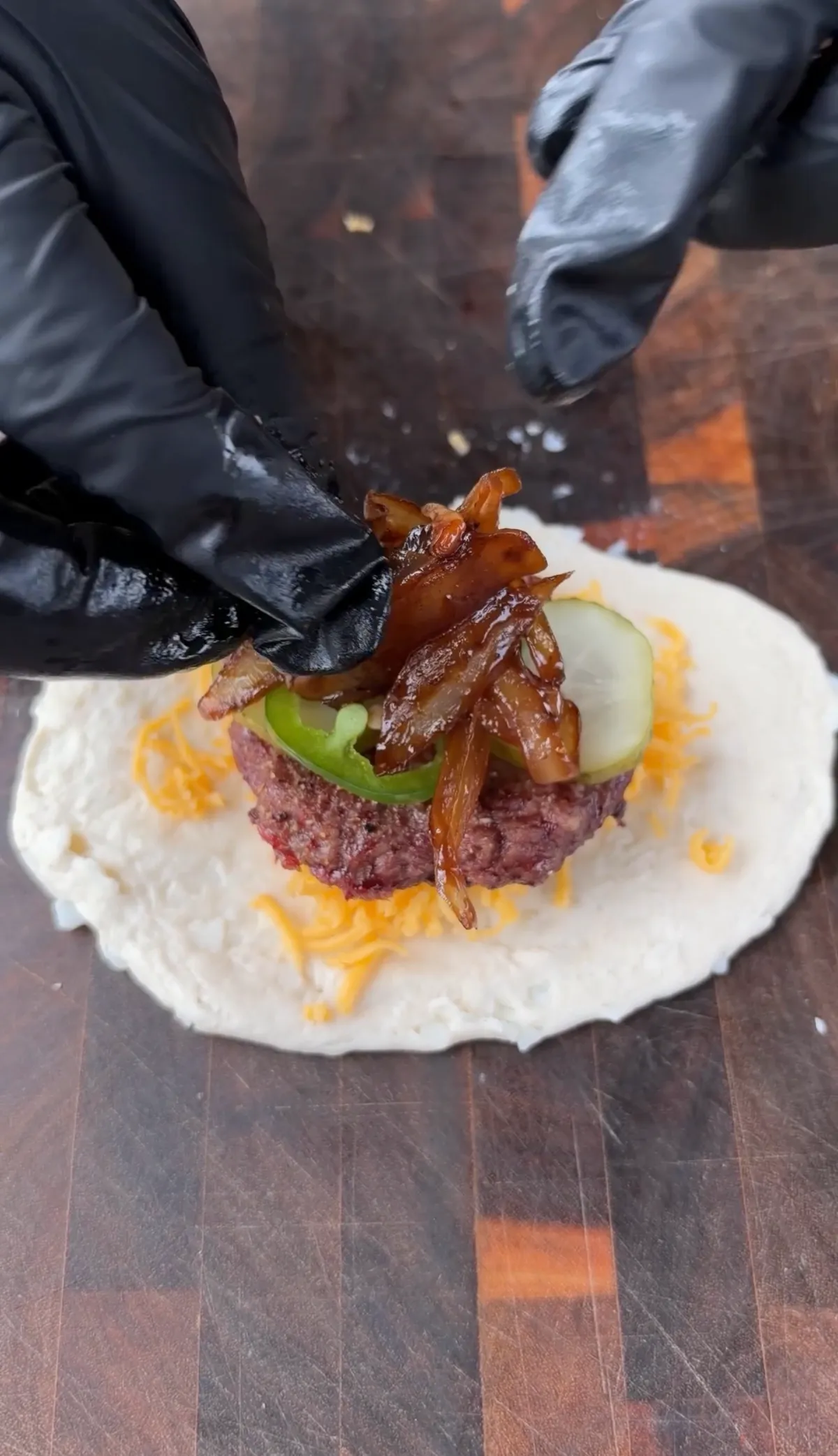 Hands layering caramelized onions onto smoked beef patty with cheddar, jalapeños, and pickles on rolled biscuit dough