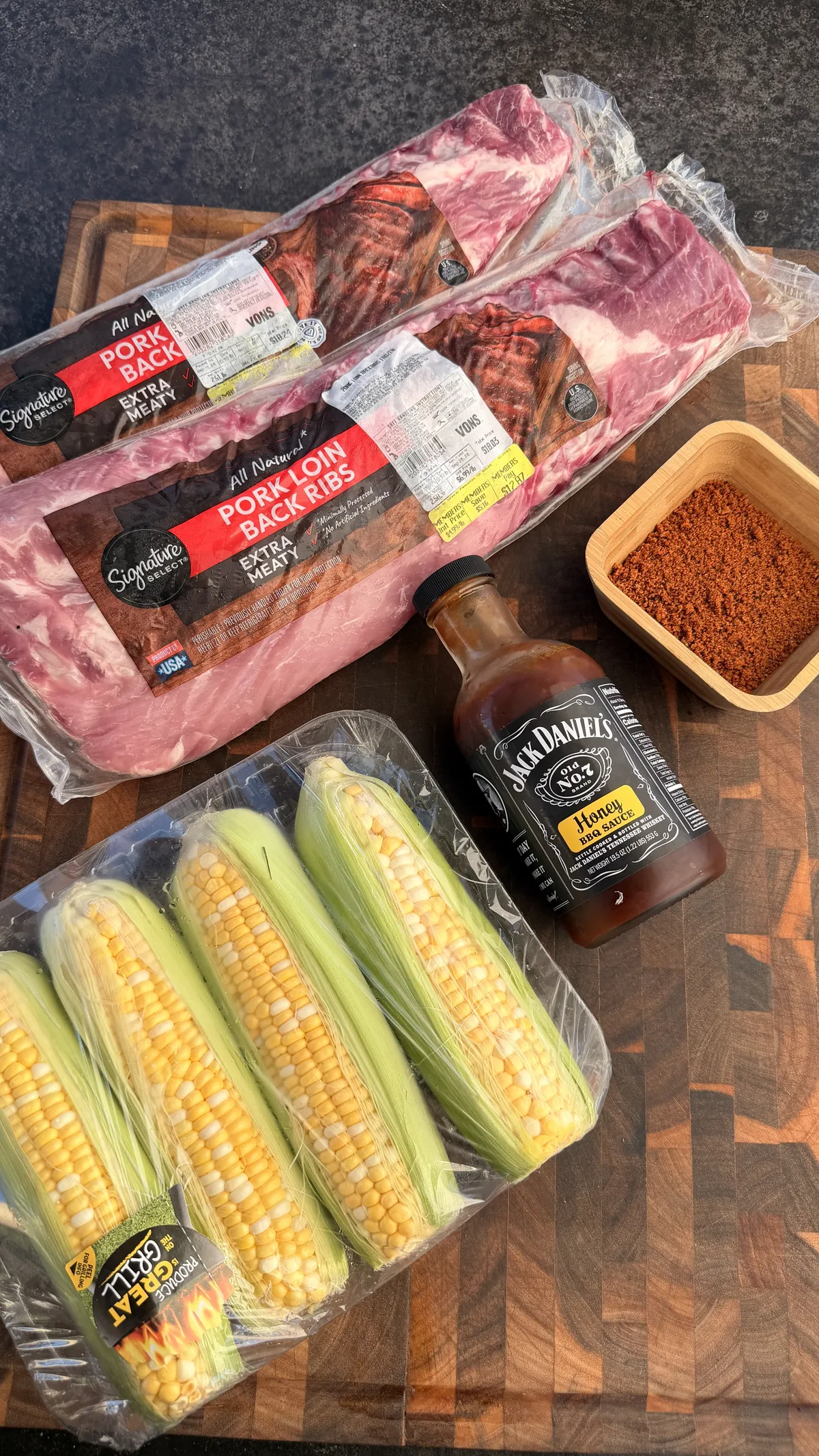 Raw baby back ribs, Jack Daniel's honey BBQ sauce, chipotle BBQ rub, and ears of corn on a cutting board