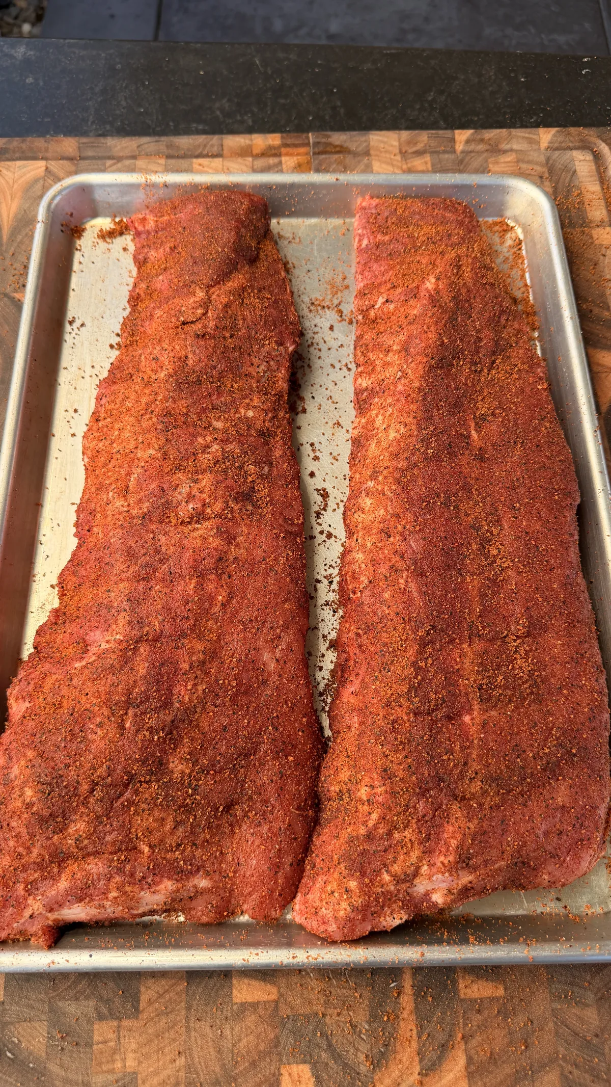 Two racks of baby back ribs generously coated in chipotle BBQ rub on a baking sheet, ready to smoke