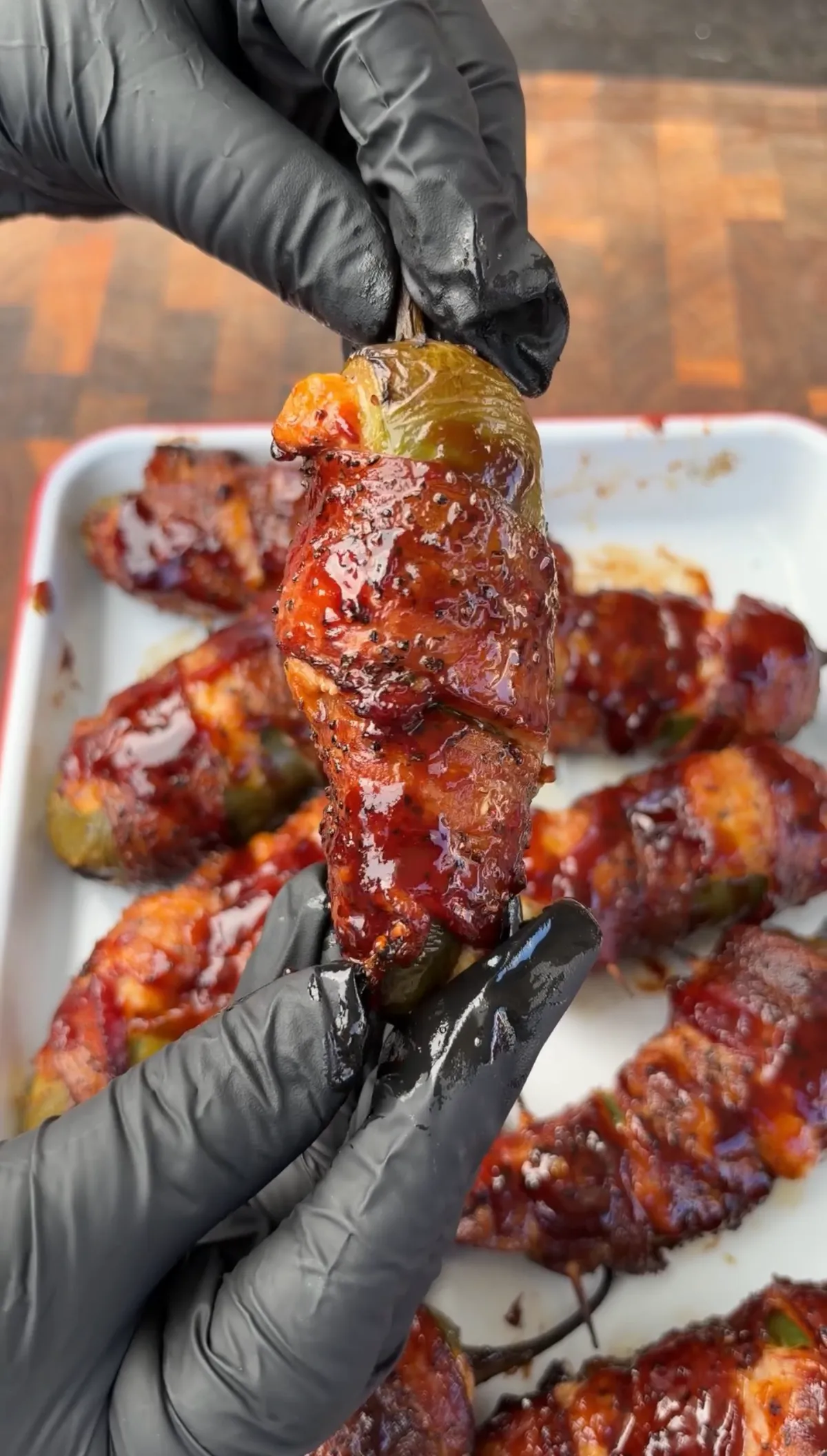 Gloved hands holding a bacon-wrapped smoked brisket-stuffed jalapeño boat glazed with sticky BBQ sauce over a tray of more