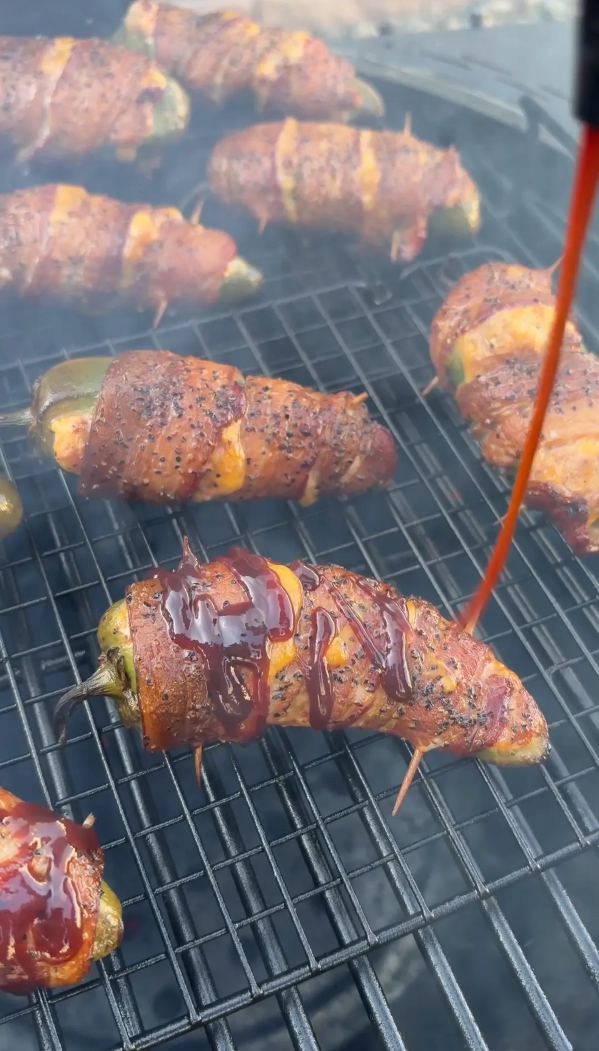 Bacon-wrapped brisket-stuffed jalapeño boats on smoker grate being glazed with sticky BBQ sauce