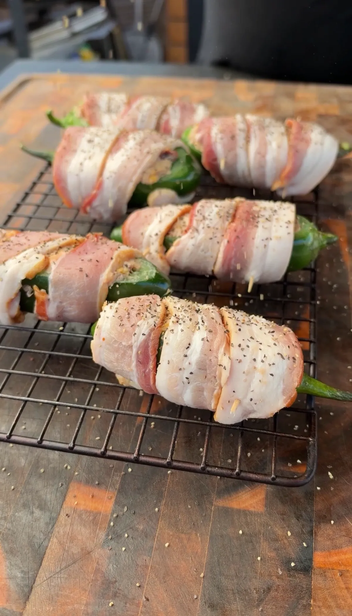 Thick-cut bacon-wrapped brisket-stuffed jalapeño boats on a wire rack, sprinkled with cracked black pepper, ready to smoke