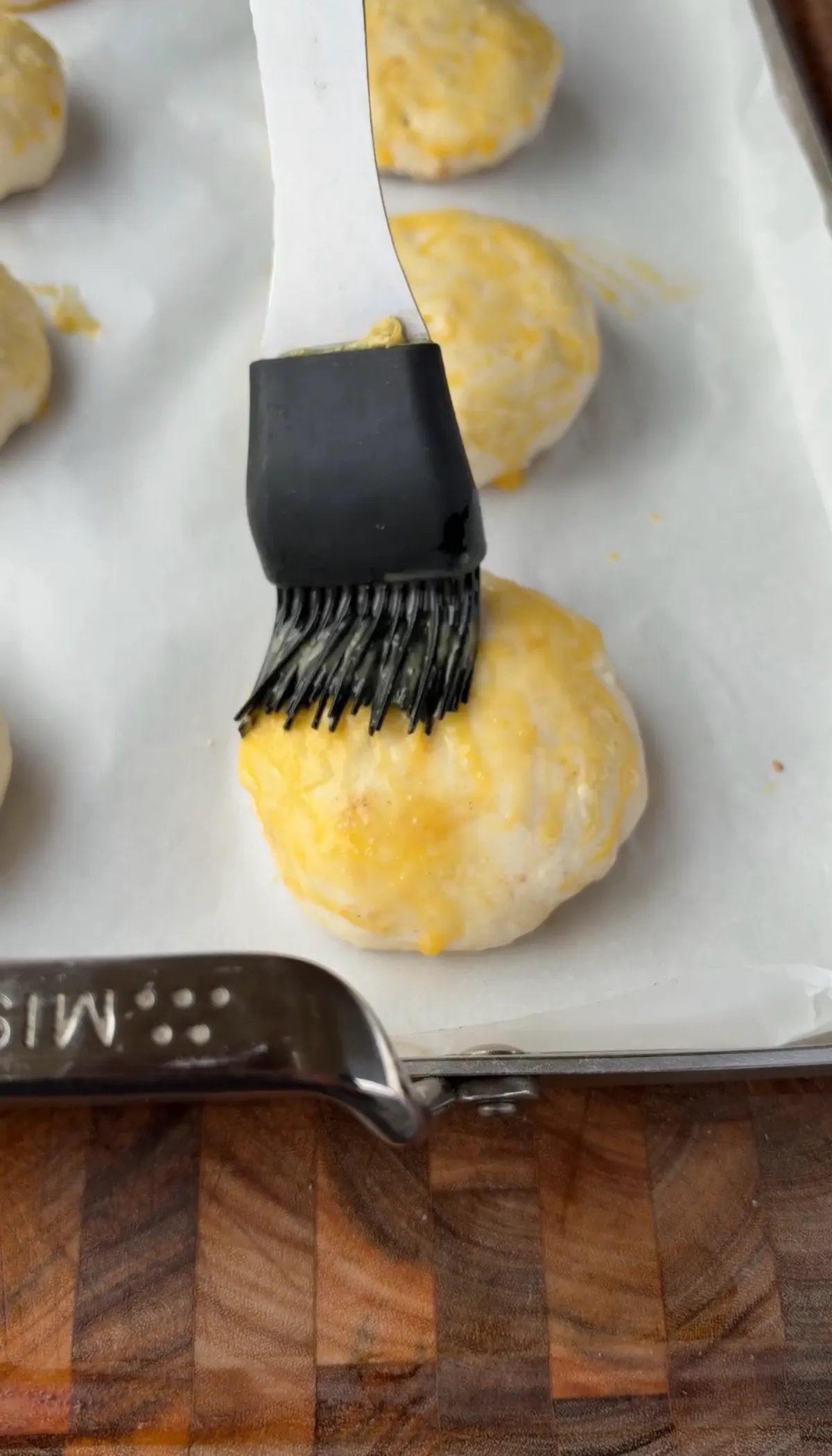 Silicone pastry brush applying egg wash to assembled smoked spicy cheeseburger bombs on parchment-lined baking sheet