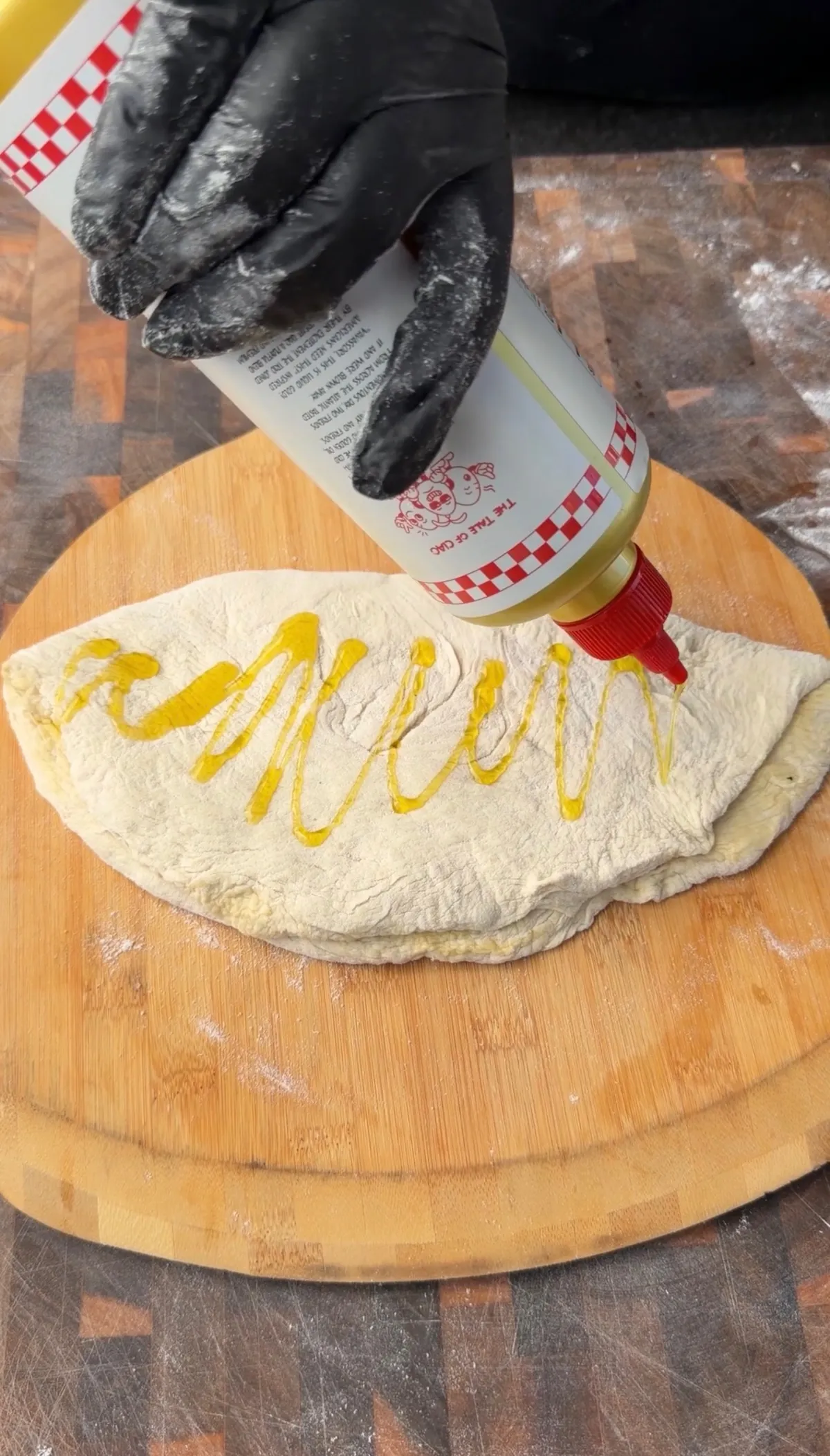 Black-gloved hand drizzling olive oil onto folded pizza dough on a wooden cutting board