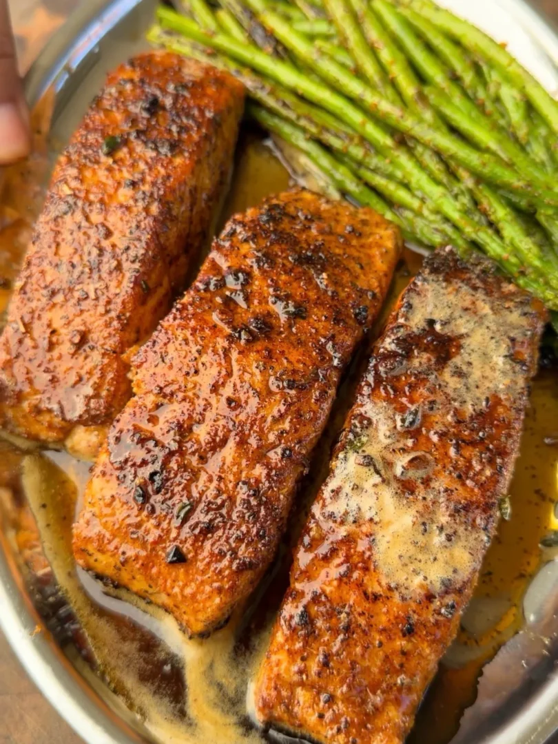 Three butter-basted Cajun-seasoned pan-seared salmon portions with thyme-infused butter and charred asparagus on a skillet
