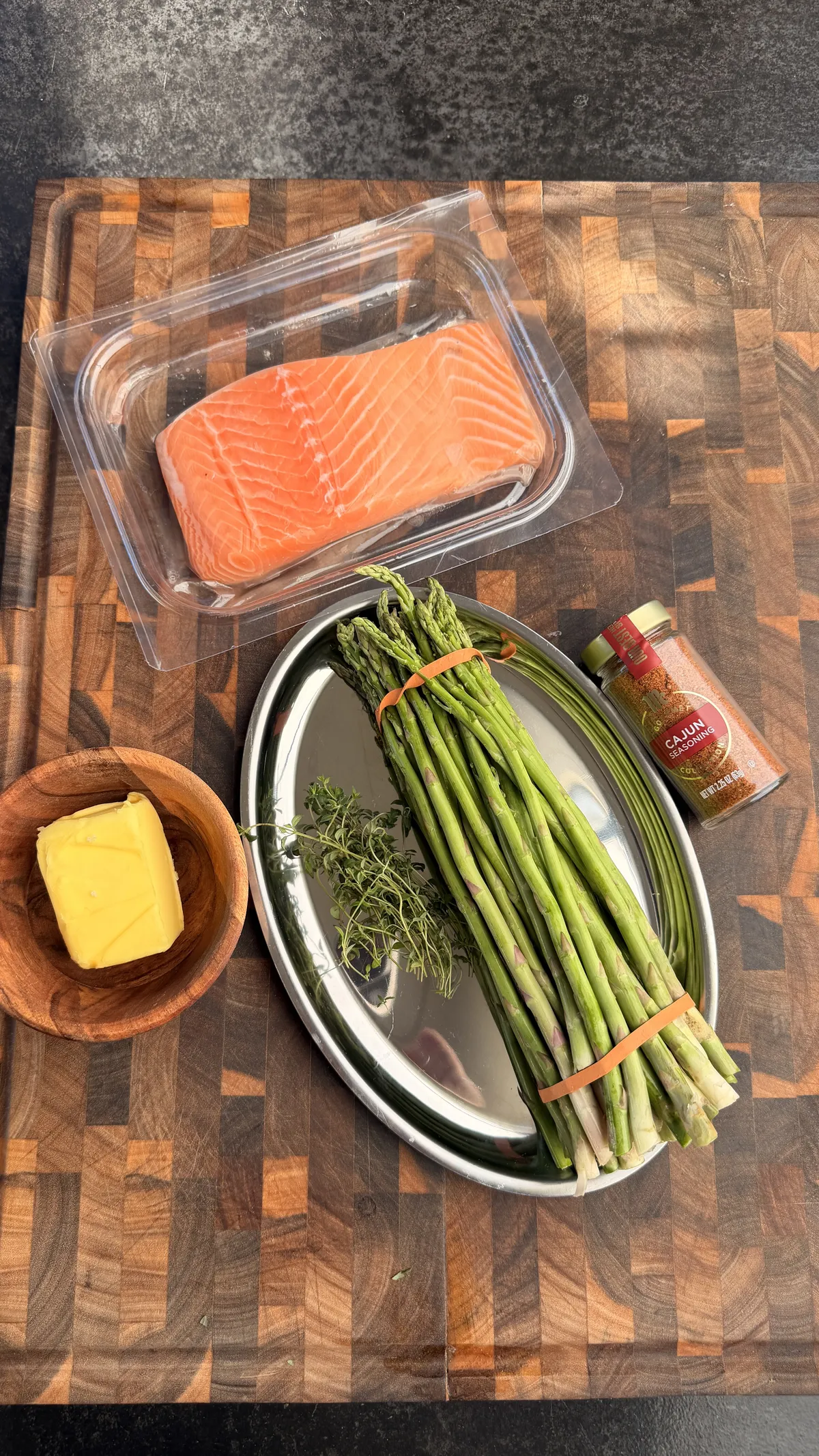 Raw salmon fillet, fresh asparagus, butter, thyme sprigs, and Cajun seasoning on a wood cutting board