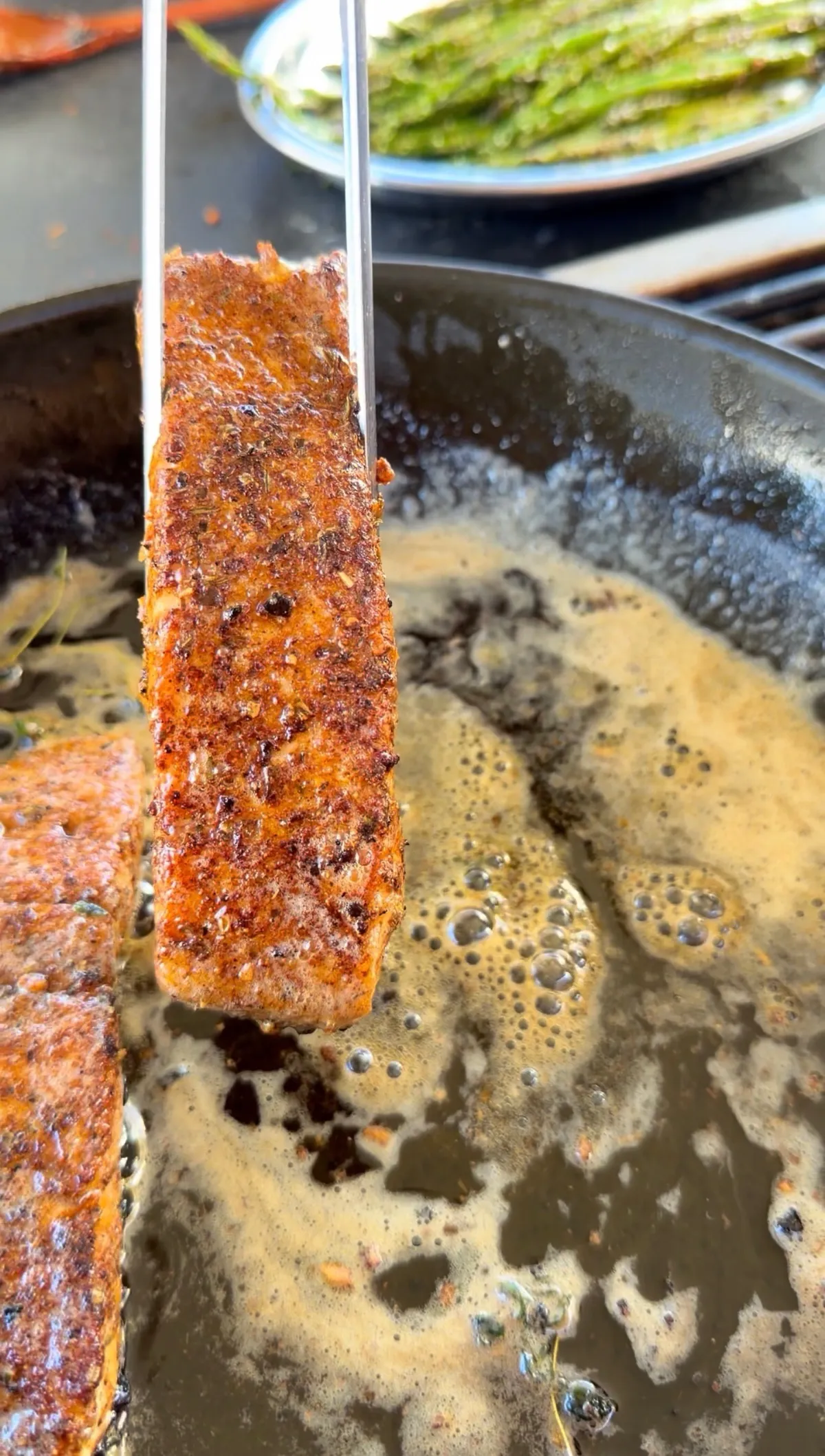 Cajun-seasoned salmon held by tongs on its side in a buttery skillet, showing a golden seared crust