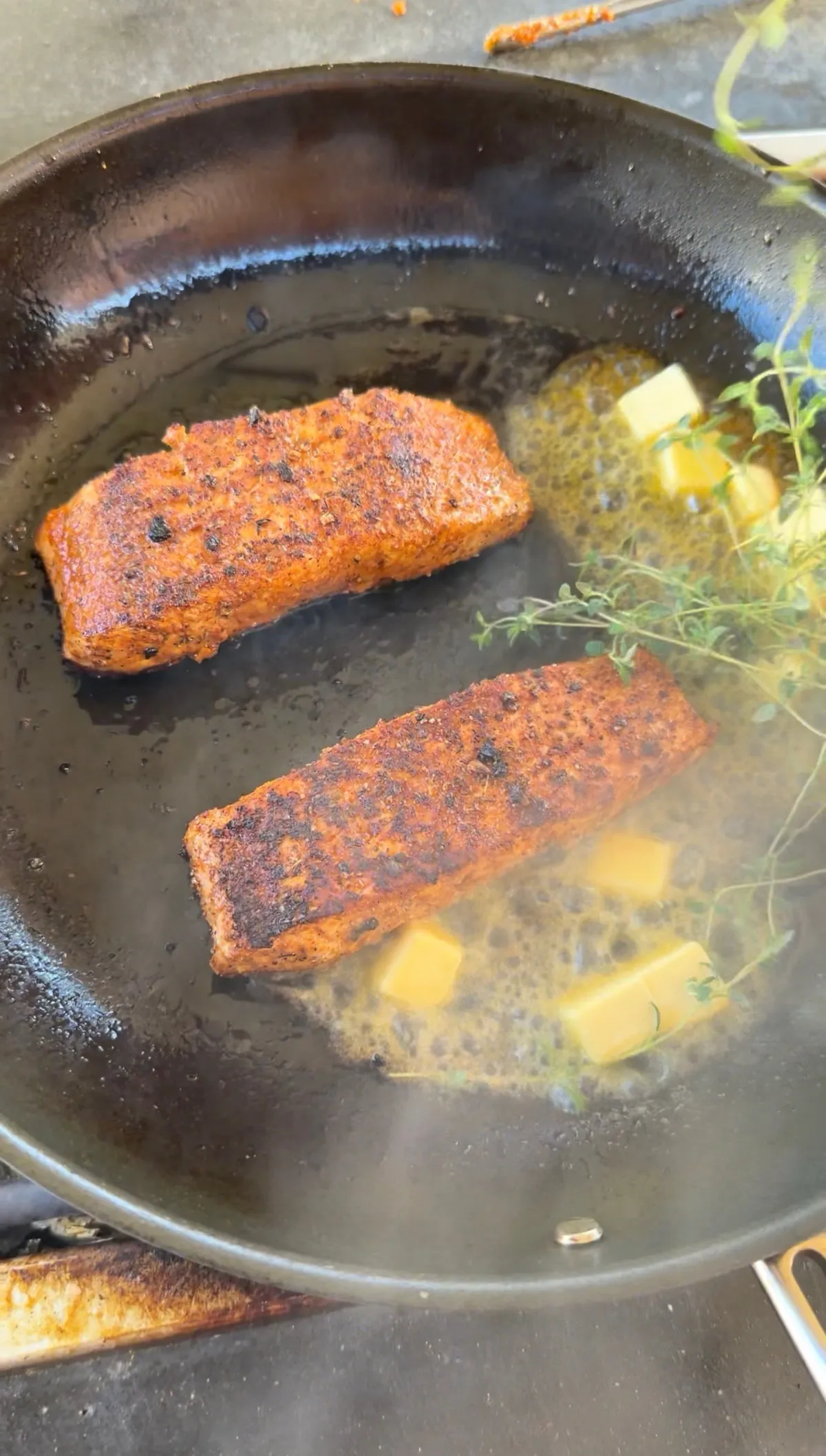 Two Cajun-seasoned salmon portions searing in a skillet with melting butter cubes and fresh thyme sprigs