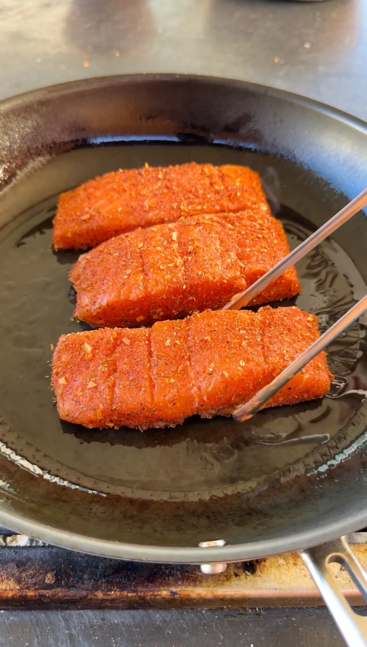 Three Cajun-seasoned salmon portions searing in a hot skillet with tongs, coated in bold red spice rub