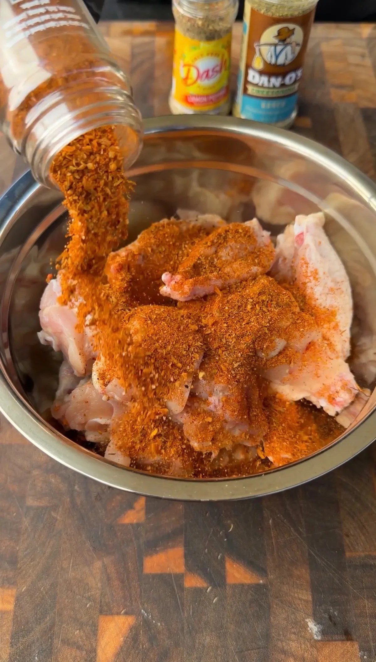 Cajun seasoning being poured from a jar over raw chicken wings in a stainless steel bowl with Dash and Dan-O's seasoning in background
