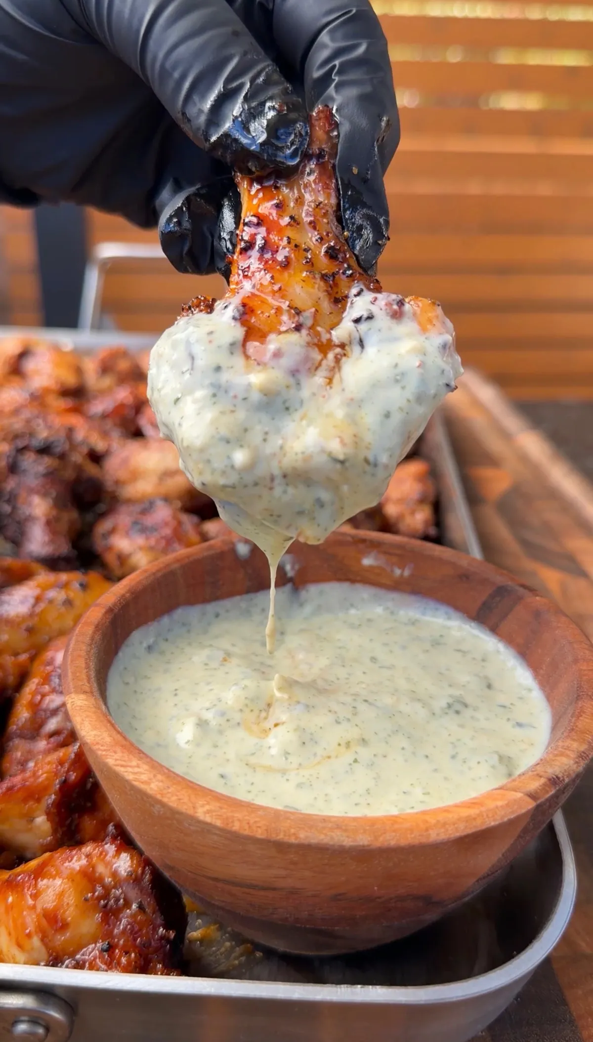 Gloved hand dipping a grilled Cajun wing into a wooden bowl of jalapeño honey butter sauce, dripping over a tray of three-way wings