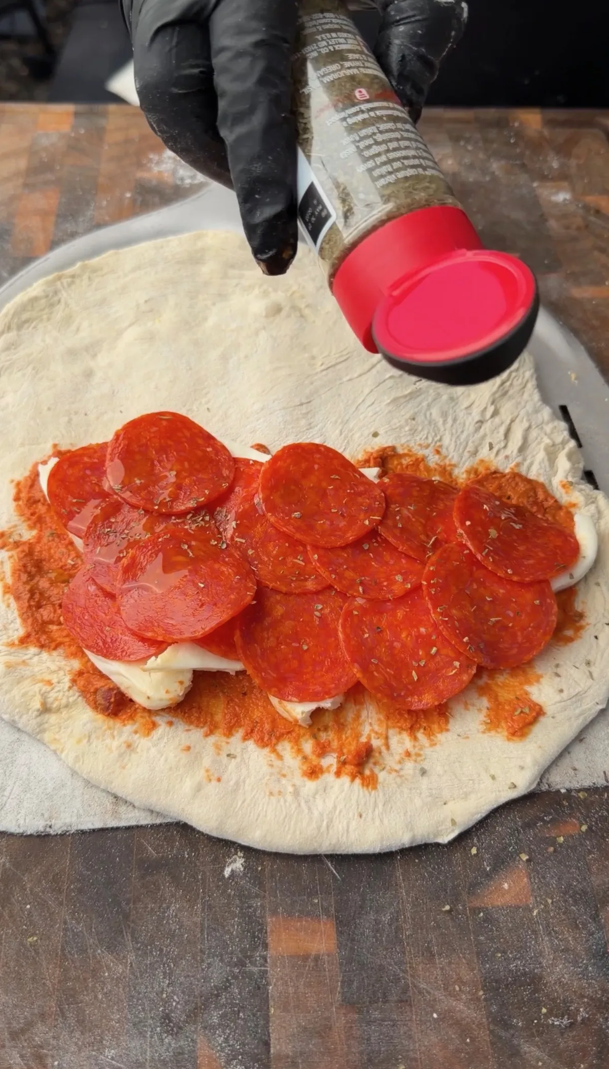 Hands in black gloves sprinkling Italian seasoning over pepperoni and fresh mozzarella on stretched pizza dough